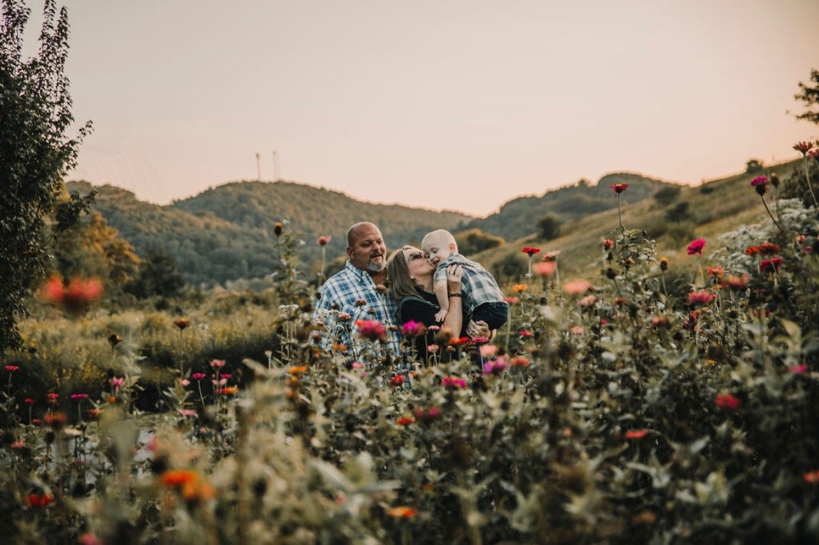A family of three in a flower field, with hills in the background, at sunset.