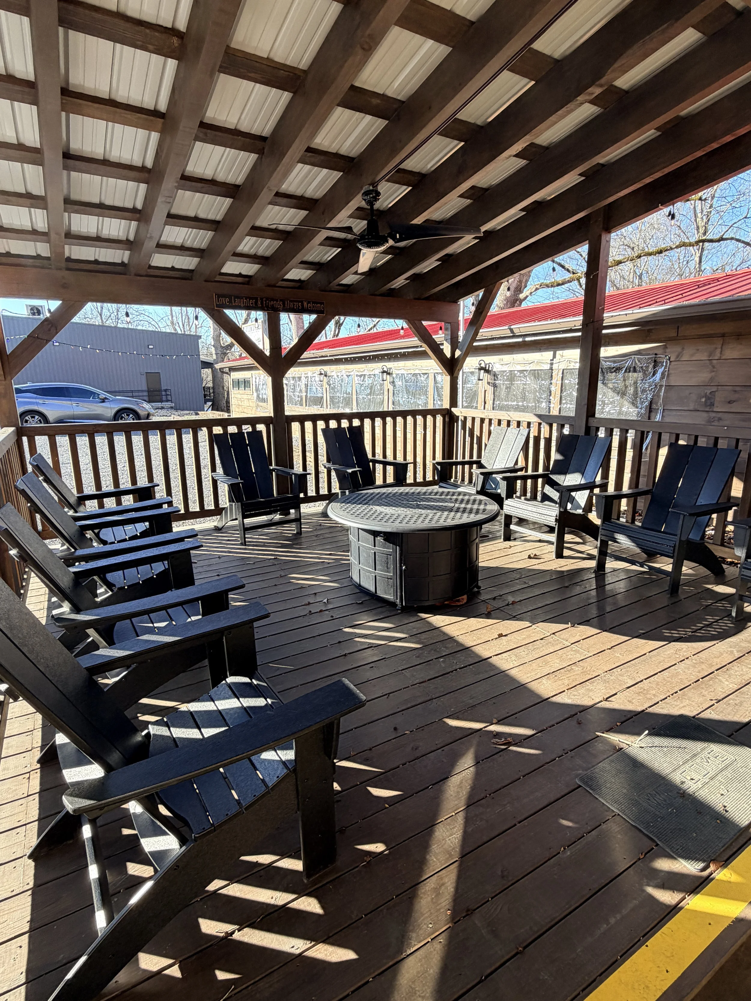 Covered wooden deck with multiple black Adirondack chairs arranged in a circle around a fire pit, under a wooden roof with a ceiling fan, during daytime.