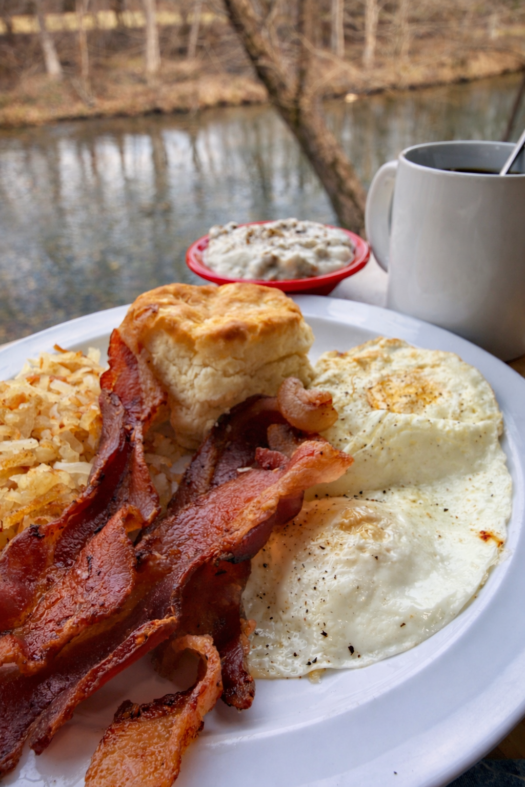 Breakfast plate with bacon, biscuits and gravy, hash browns, and fried eggs, set outdoors near a river with trees in the background.