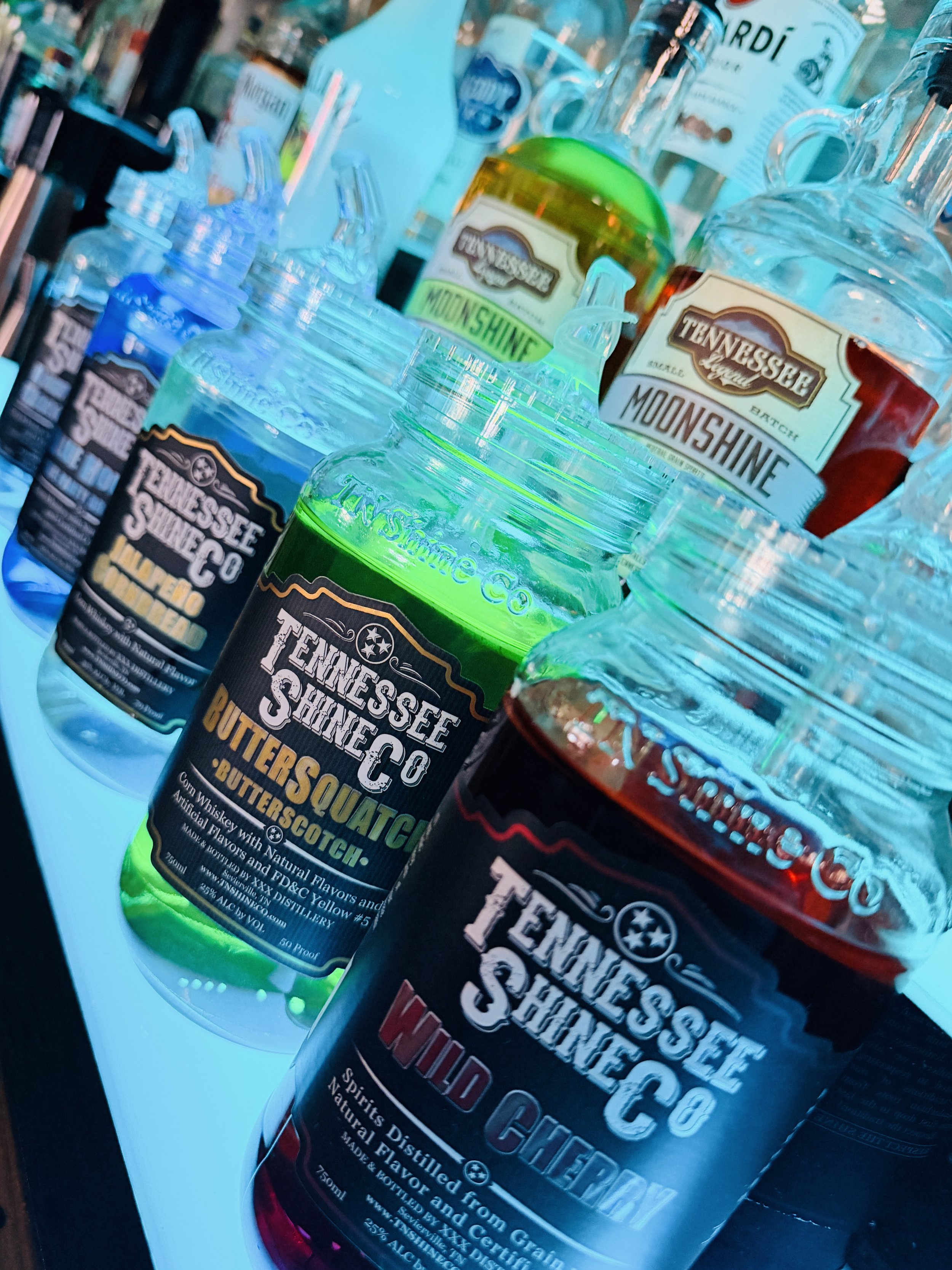 Display of colorful bottles of Tennessee Shine Co whiskey, including flavors like Butterscotch and Wild Cherry, on a lit-up surface at a store or bar.