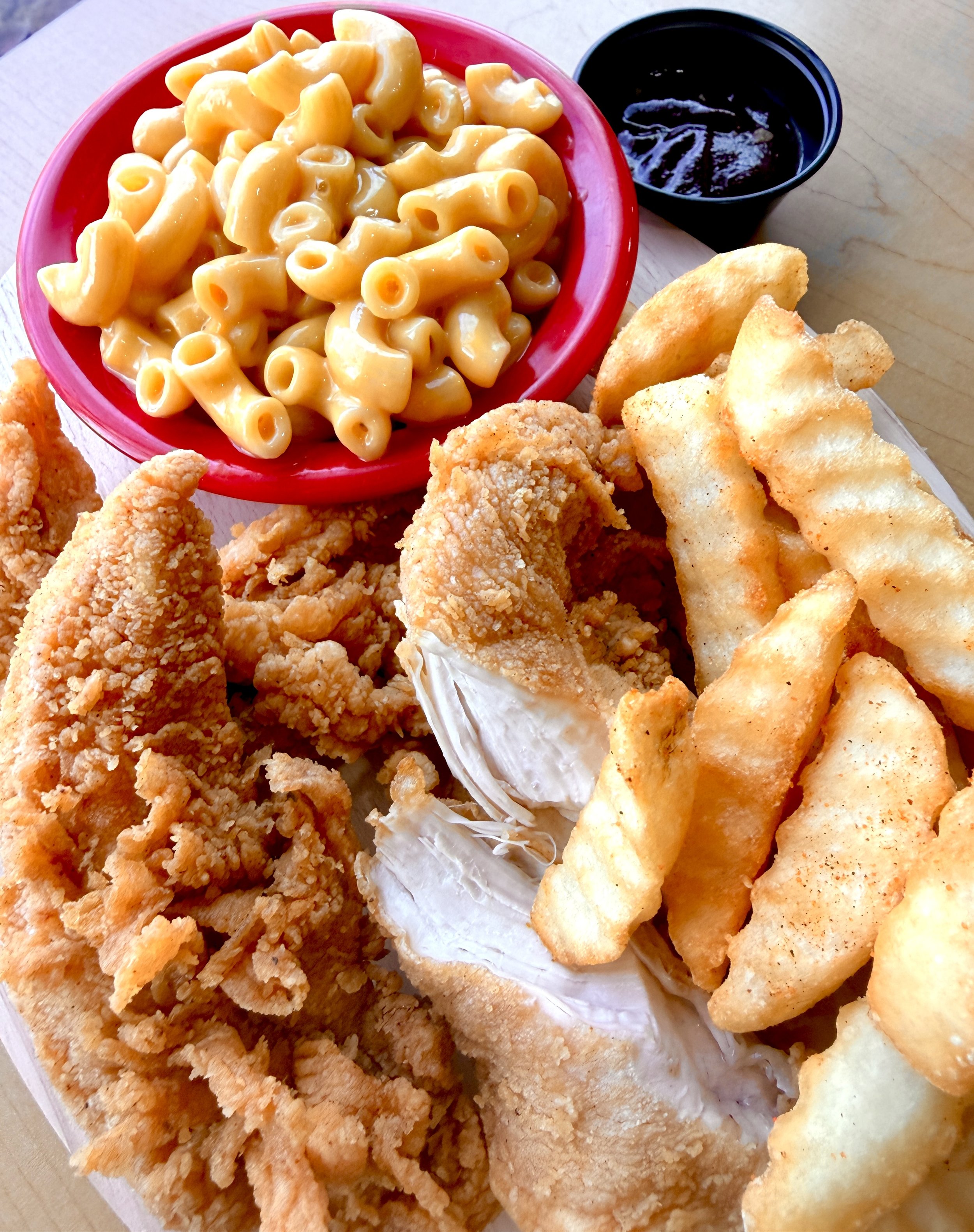 Fried chicken tenders, French fries, fried fish fillet, macaroni and cheese in a red bowl, and a small cup of dark sauce on a light wooden surface.