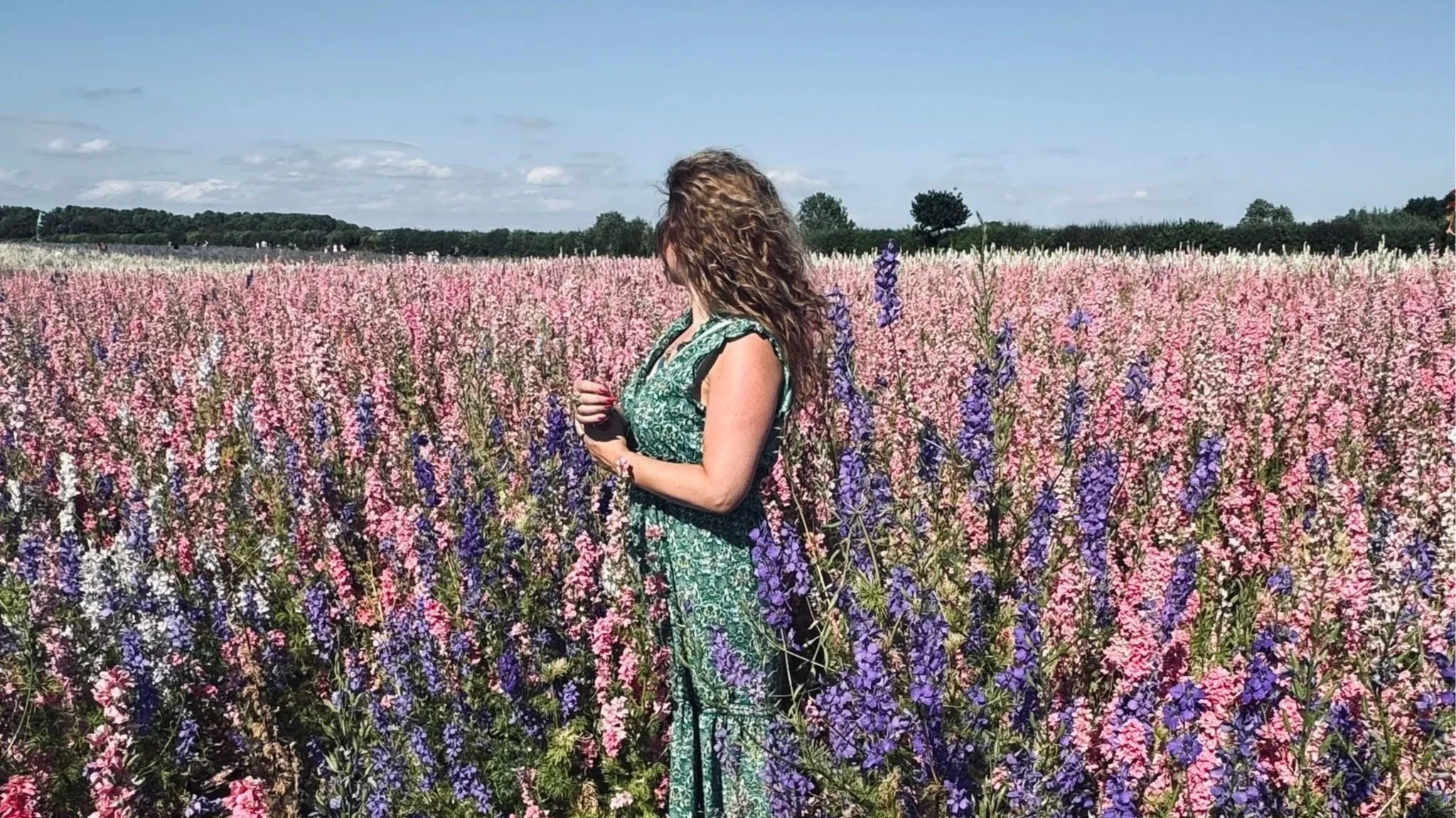 A woman in a green dress, Gemma Pinfield-Thomas, standing in a flower field with pink, purple, and white delphinium flowers, looking away from the camera.