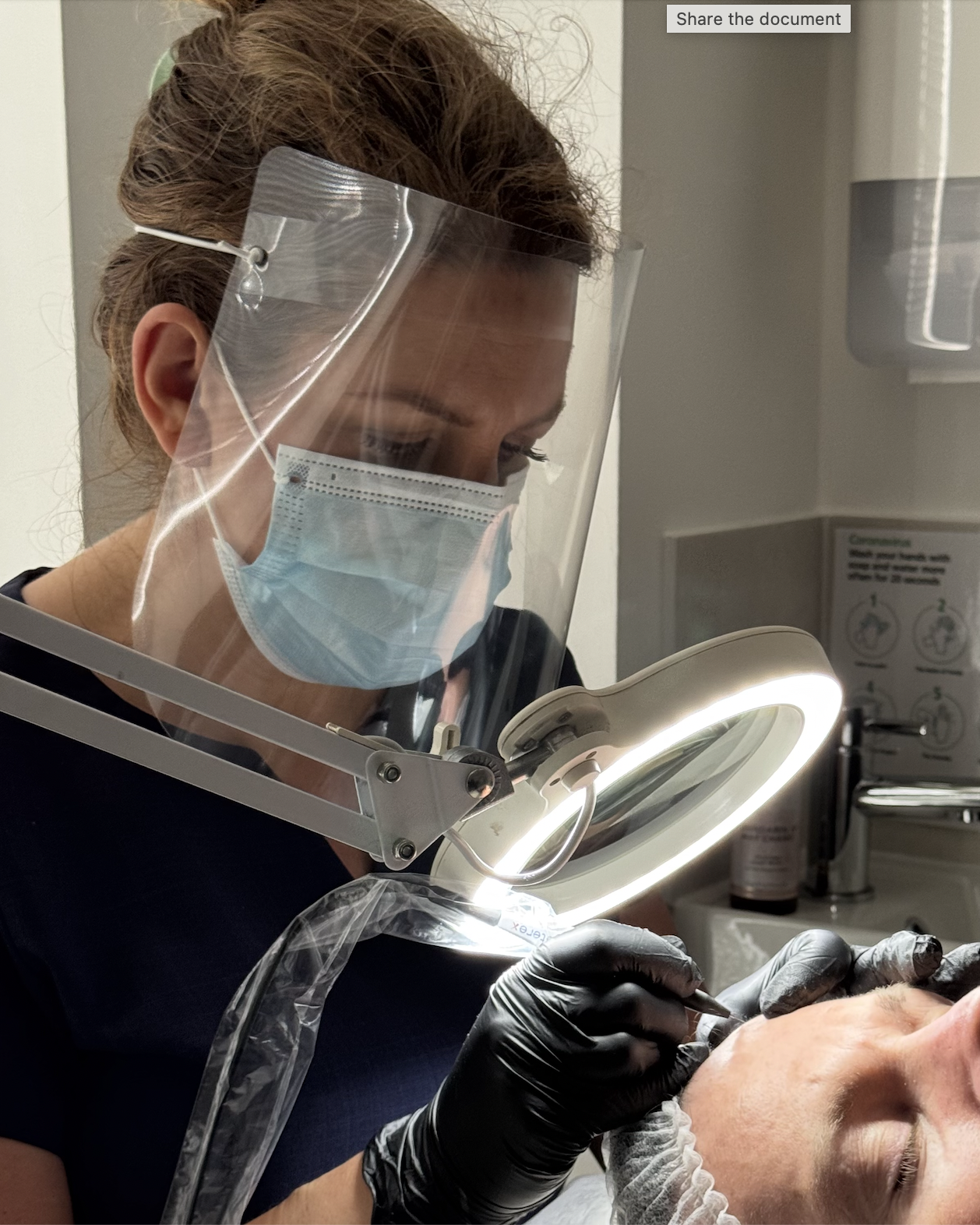 Advanced electrologist Gemma Pinfield-Thomas wearing a face mask, face shield, and black gloves is examining a cyst on a patient's forehead under a bright ring light in a clinical setting.