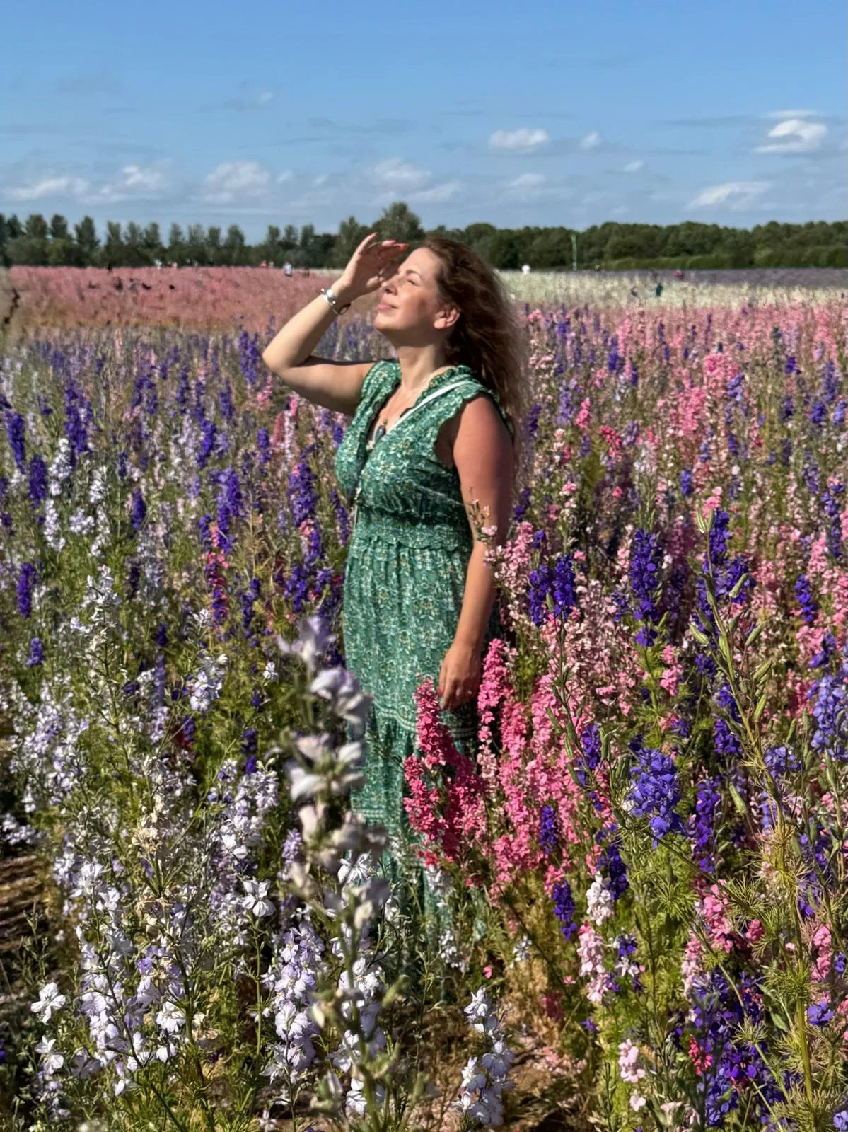 A woman in a green dress, Gemma Pinfield-Thomas, standing in a colourful flower field of pink, purple and white delphinium flowers, her eyes closed and one hand shielding her eyes from the sun, under a blue sky with scattered clouds.