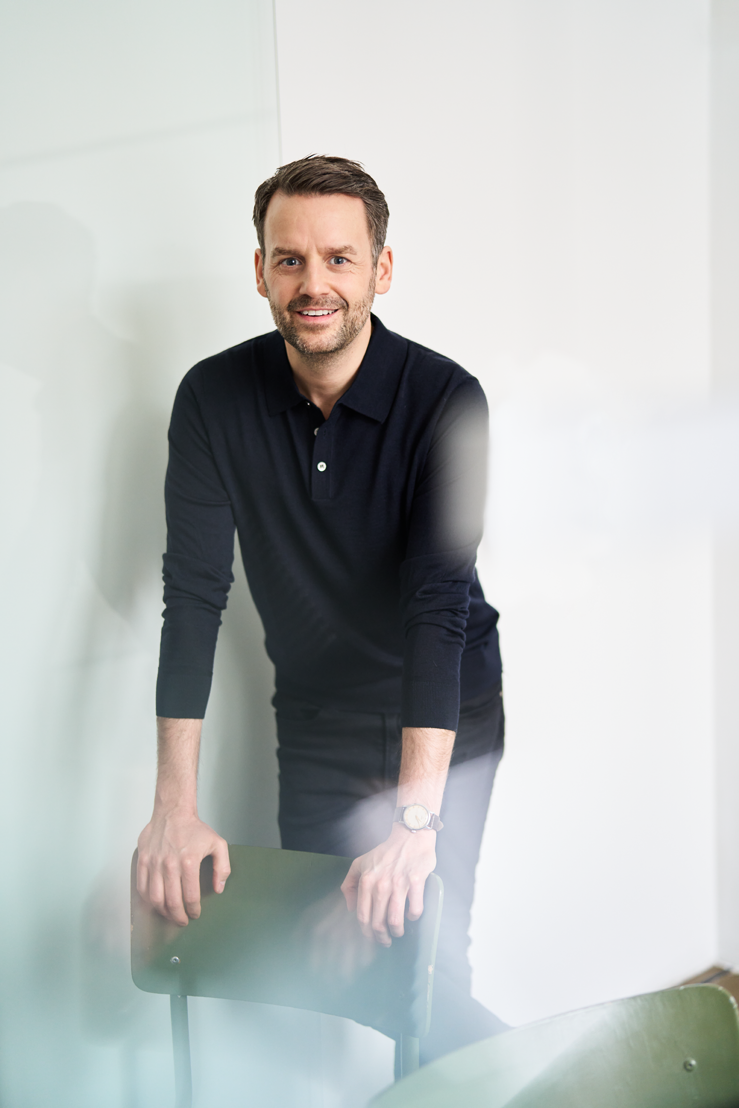 Ein Porträt von Christoph P. Deneke, wie er in einem dunkelblauen Sweatshirt, Schwarzer Jeans auf einen grünen Stuhl gestützt ist. Foto geschossen von dem Fotografen Benne Ochs.
