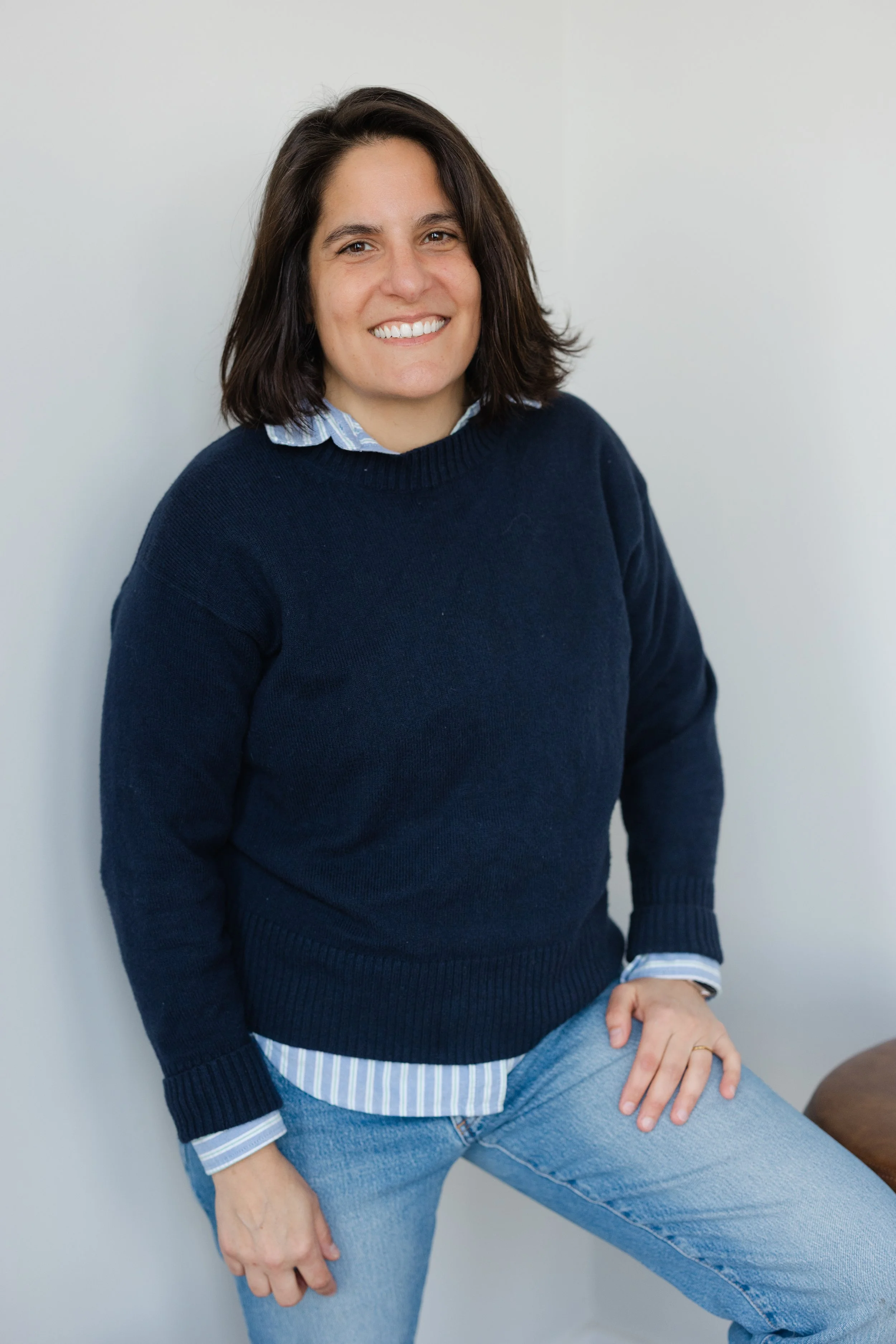 A woman with dark brown hair and a wide smile, wearing a navy blue sweater over a striped collared shirt, standing against a plain white wall.
