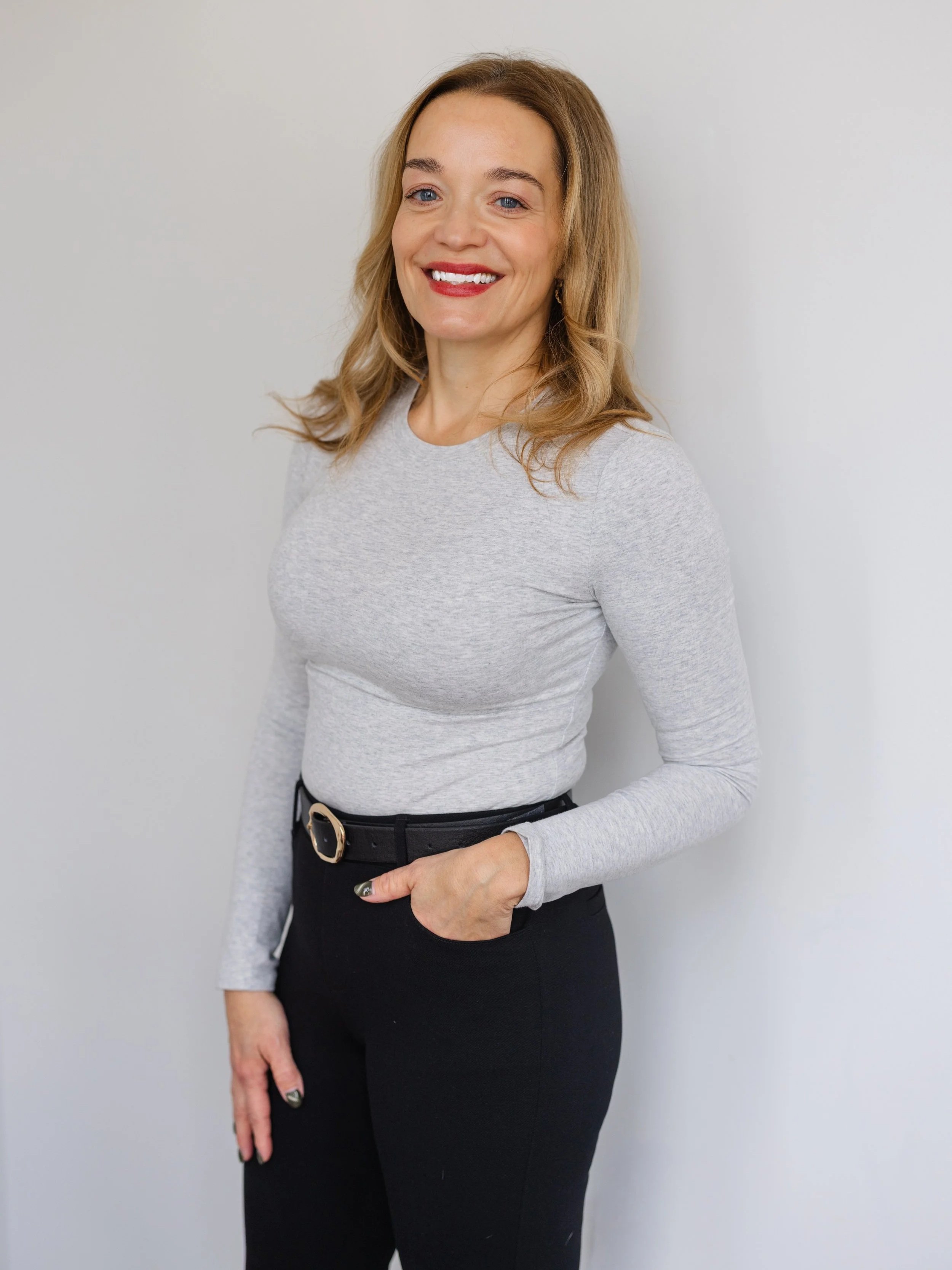 A smiling woman with blonde hair wearing a light gray long sleeve top and black pants standing against a light gray wall.