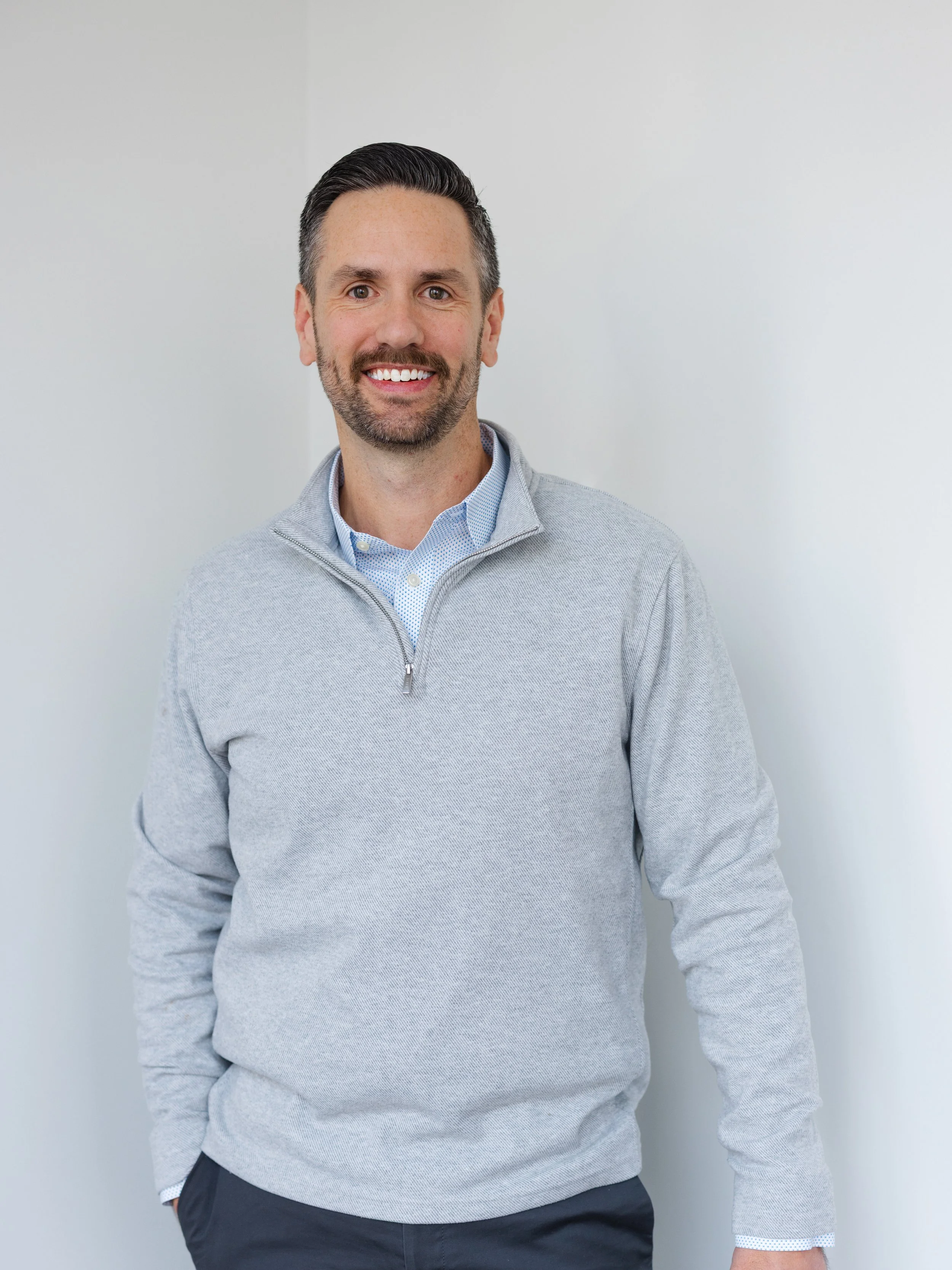 Portrait of a smiling man with dark hair and beard, wearing a light gray zip-up sweater and a blue collared shirt, standing against a plain light-colored wall.