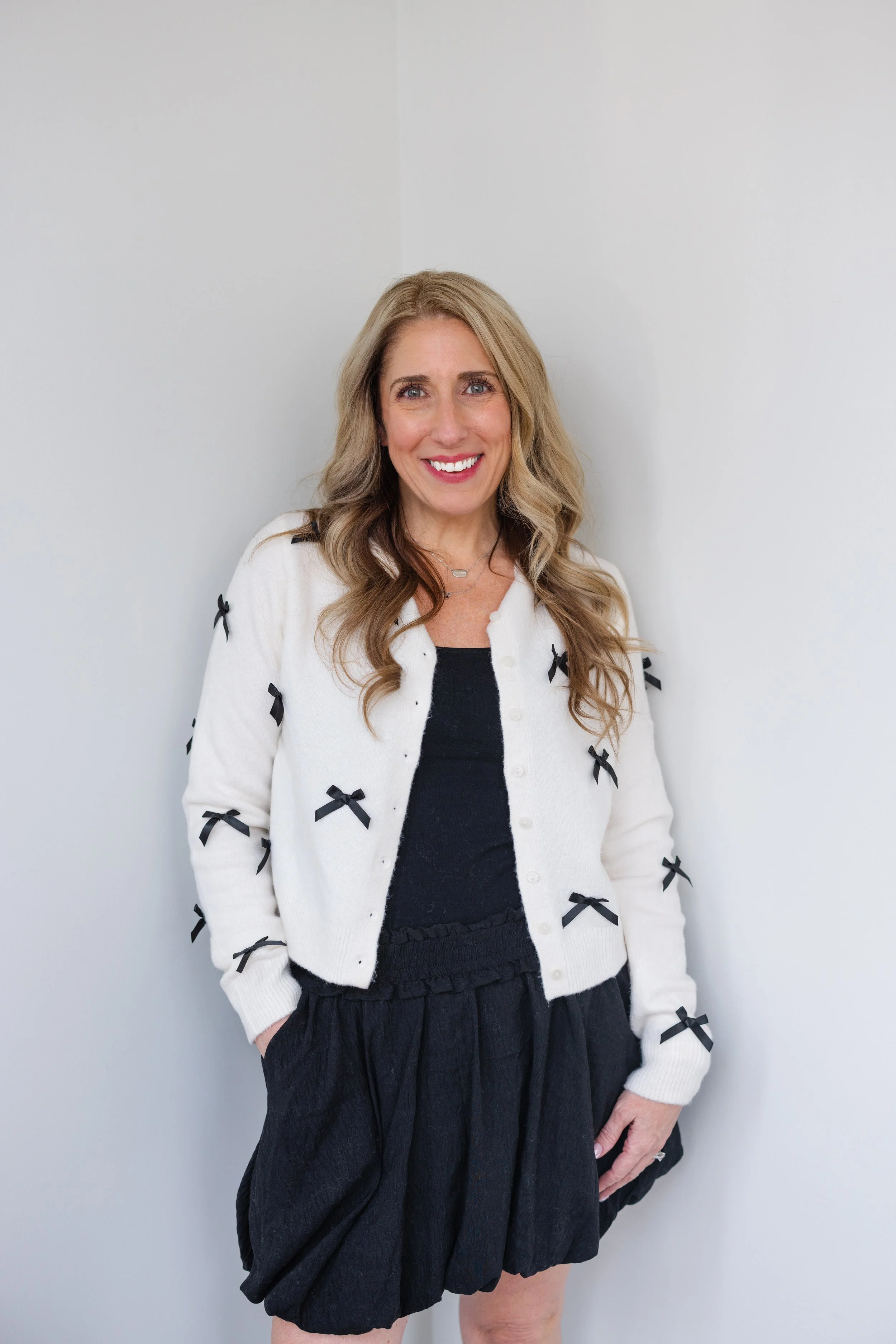 A woman with long, wavy blonde hair is smiling and standing against a plain white wall. She is wearing a white cardigan with black bows, a black top, and black skirt.