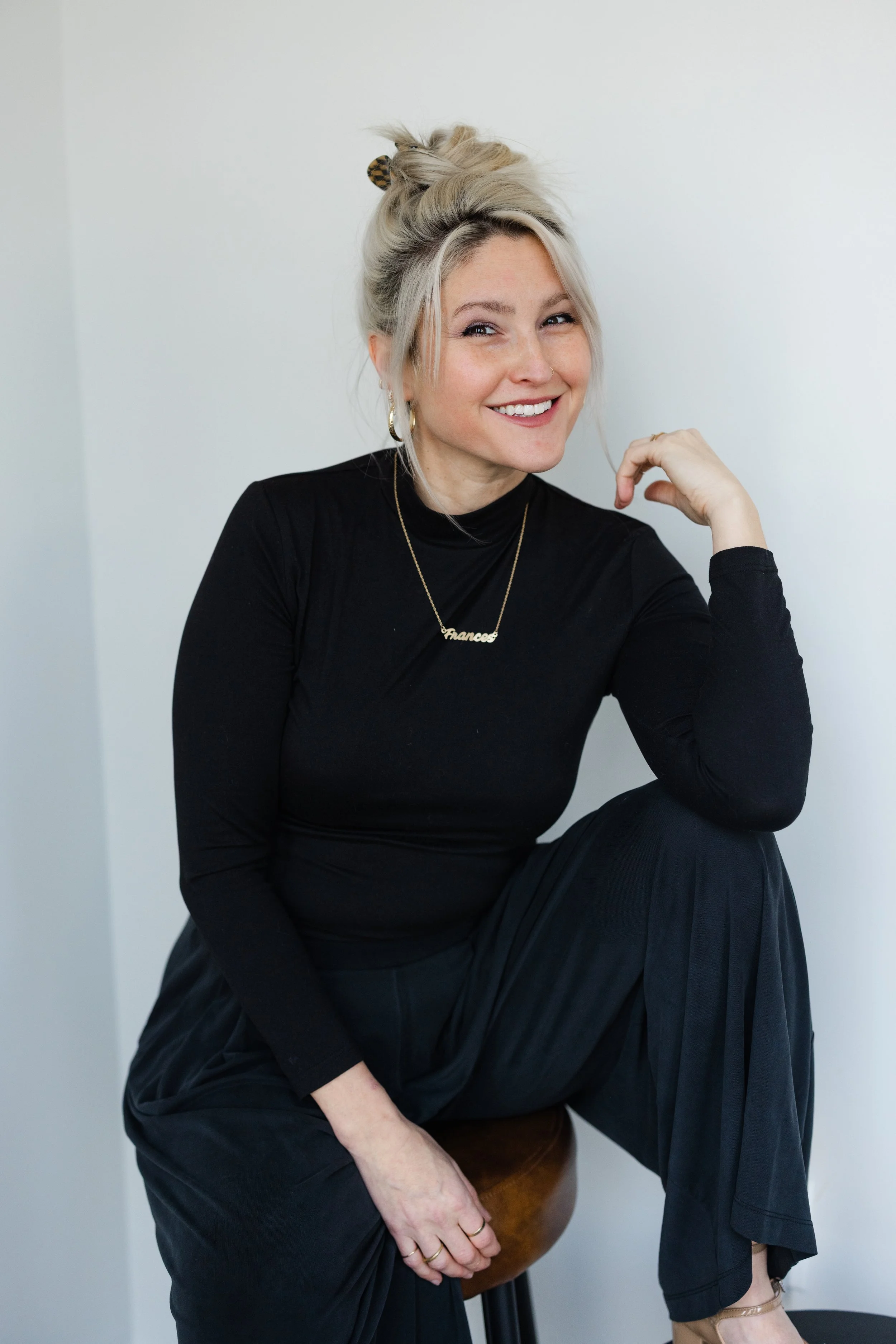 A smiling woman with blonde hair tied up, wearing a black long-sleeve top, black pants, and beige shoes, sitting on a wooden stool against a plain white wall.