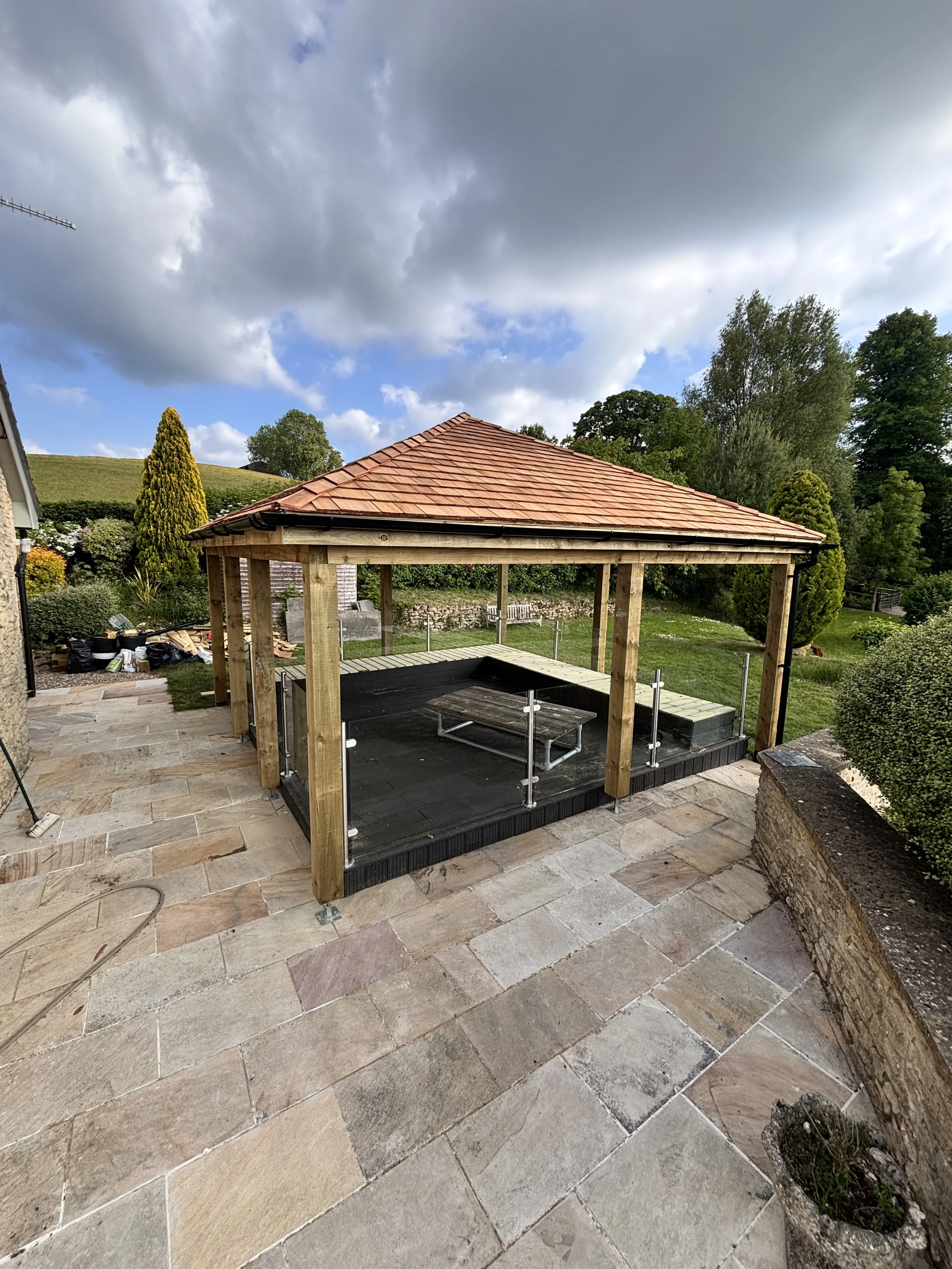 View of a backyard with a newly constructed wooden gazebo with a tiled roof, glass railing, and a small table inside, surrounded by trees and a partly cloudy sky.