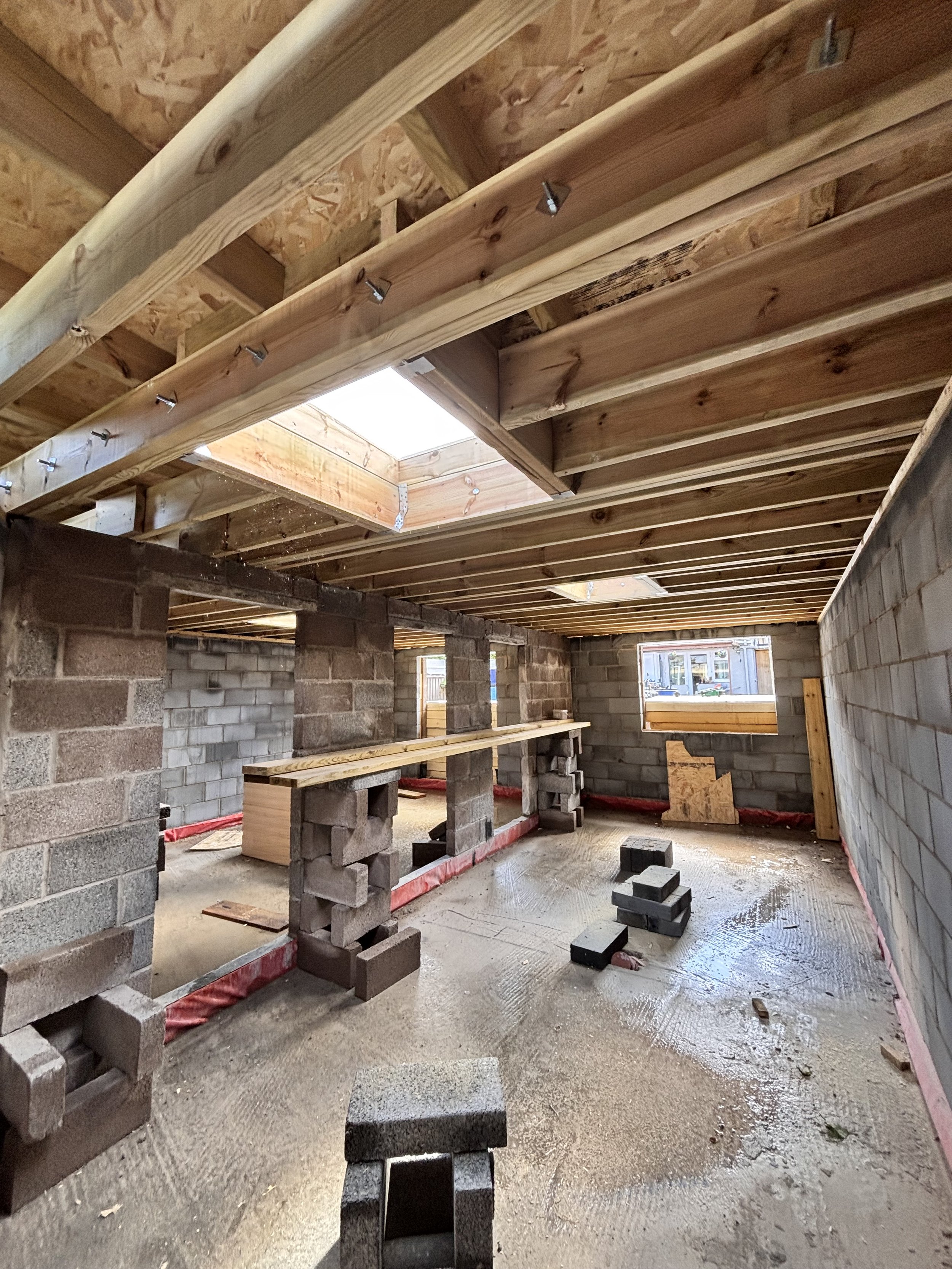 Interior view of a building under construction with brick and concrete walls, a wooden ceiling framework, and an open skylight, with construction materials and tools visible on the floor.
