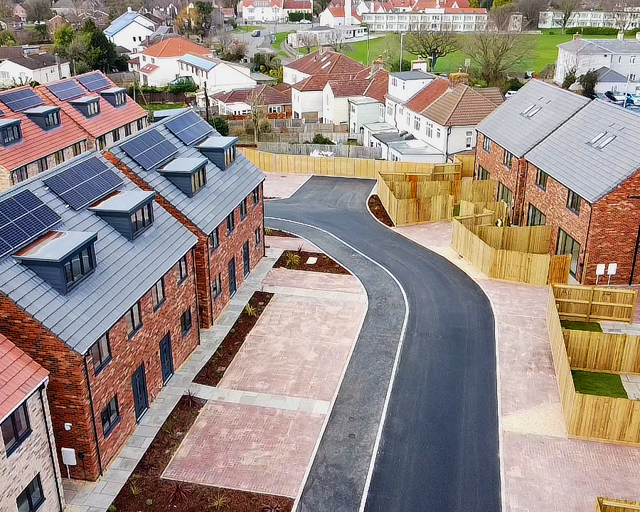 Aerial view of a new residential neighborhood with brick townhouses having solar panels on their roofs, paved walkways, driveways, and wooden fences surrounding backyards.