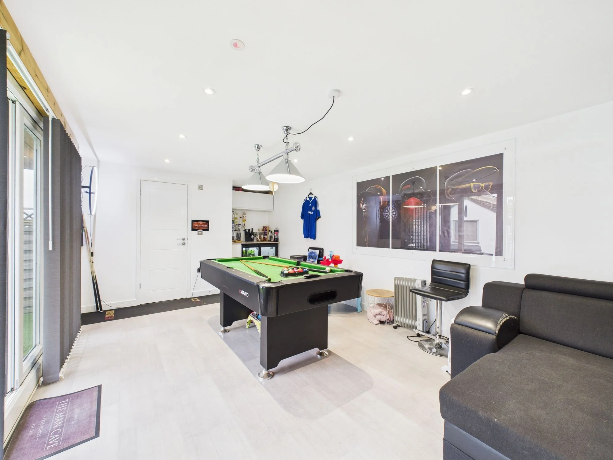 A game room with a pool table, black sofa, black chair, and bar area, featuring a large framed poster of helmets and glasses on the wall, and sliding glass doors with curtain on the left.