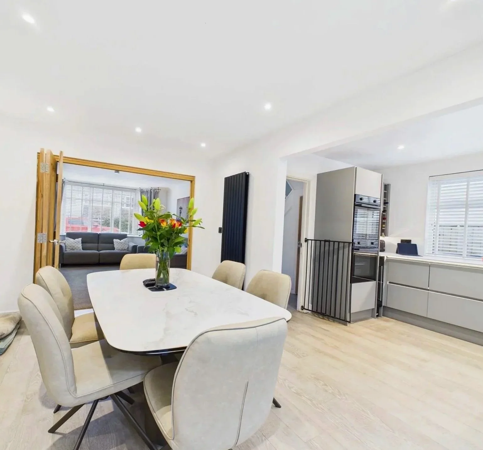 Dining area with a white marble table and eight beige chairs, a vase with colorful flowers as decoration, and a view into a living room with a gray sofa and large windows with blinds.