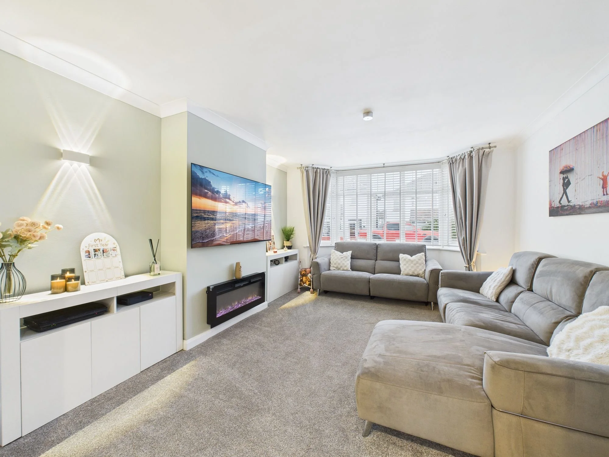 Bright living room with beige sofas, large window with white blinds and cream curtains, wall-mounted TV, electric fireplace, and decorative items on a white cabinet.