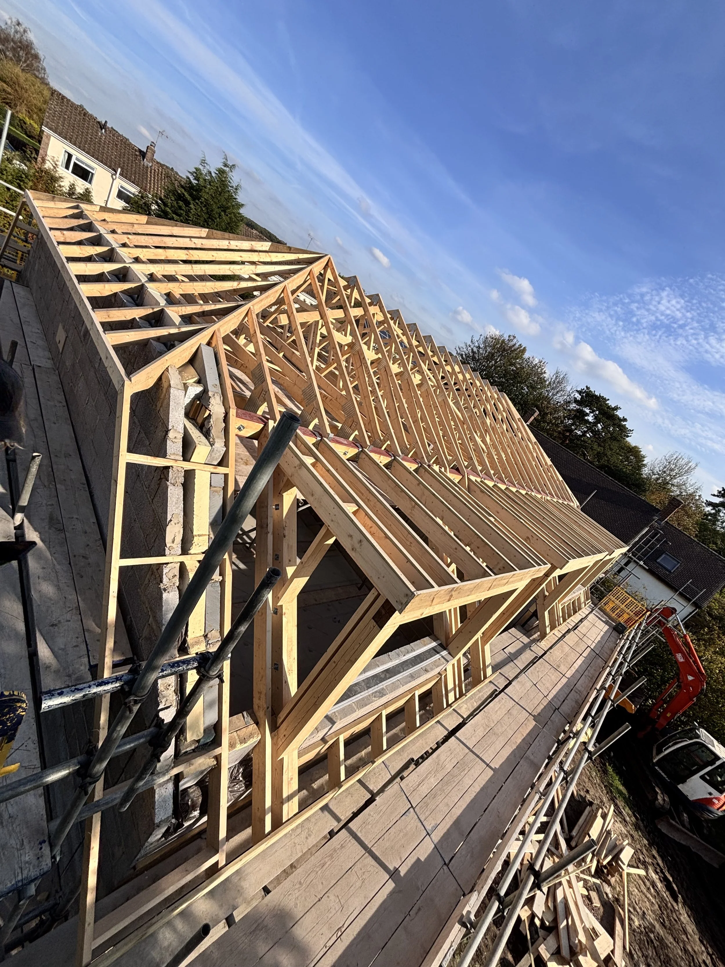 Construction site with new wooden roof framing on a building, scaffolding, and construction equipment outdoors under a blue sky with some clouds.