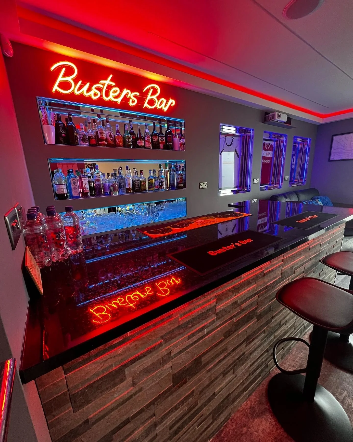 A modern bar with a illuminated red neon sign that reads 'Busters Bar.' The bar counter has a reflective surface with a neon reflection. Behind the bar, shelves are stocked with various bottles of alcohol. There are some bar stools in front of the counter and a seating area with a sofa and windows decorated with sports jerseys.