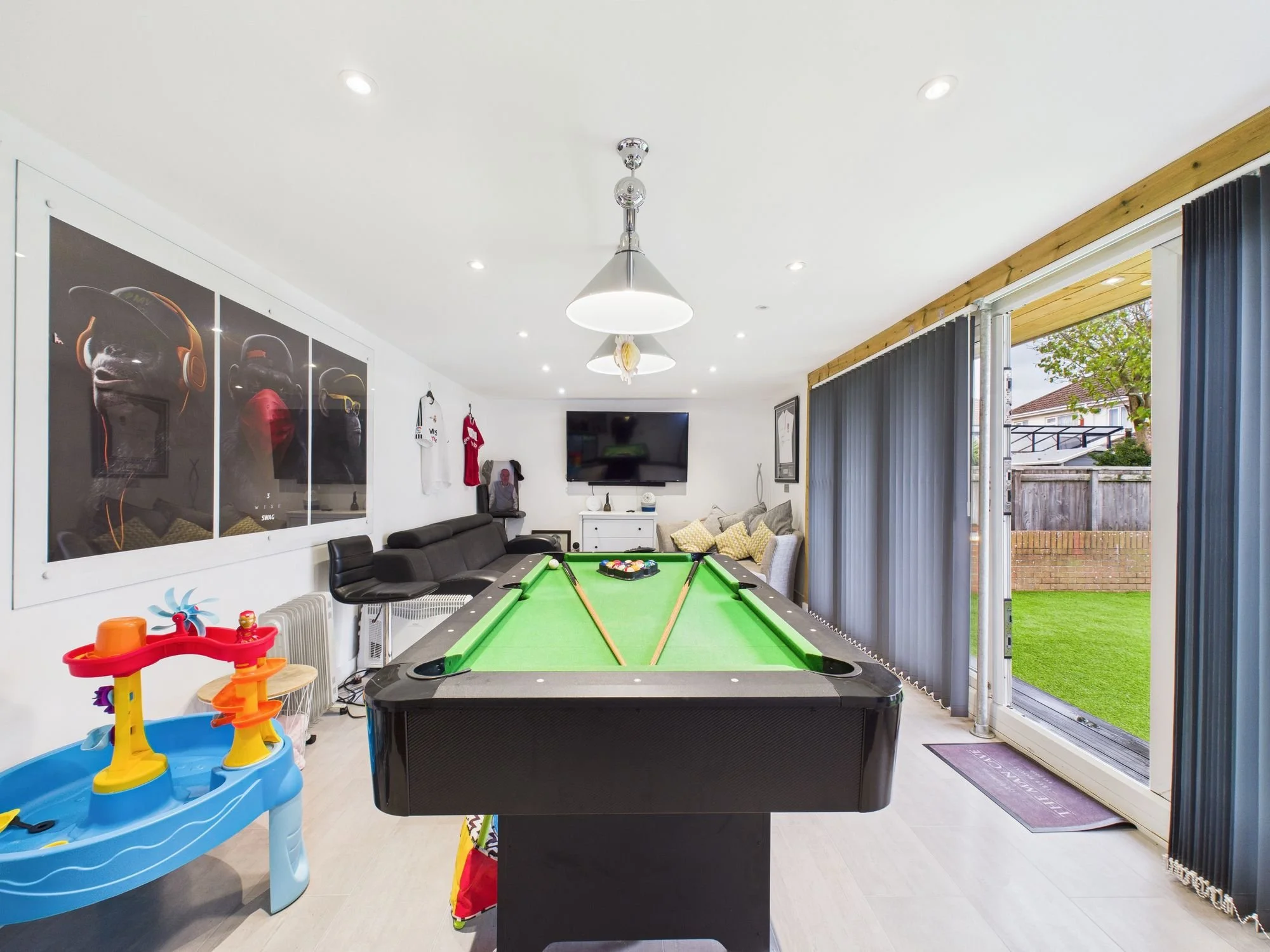 Indoor recreational room with a pool table, a large TV on the wall, a sectional sofa with pillows, children’s toy, and sliding glass door with vertical blinds leading to a backyard.
