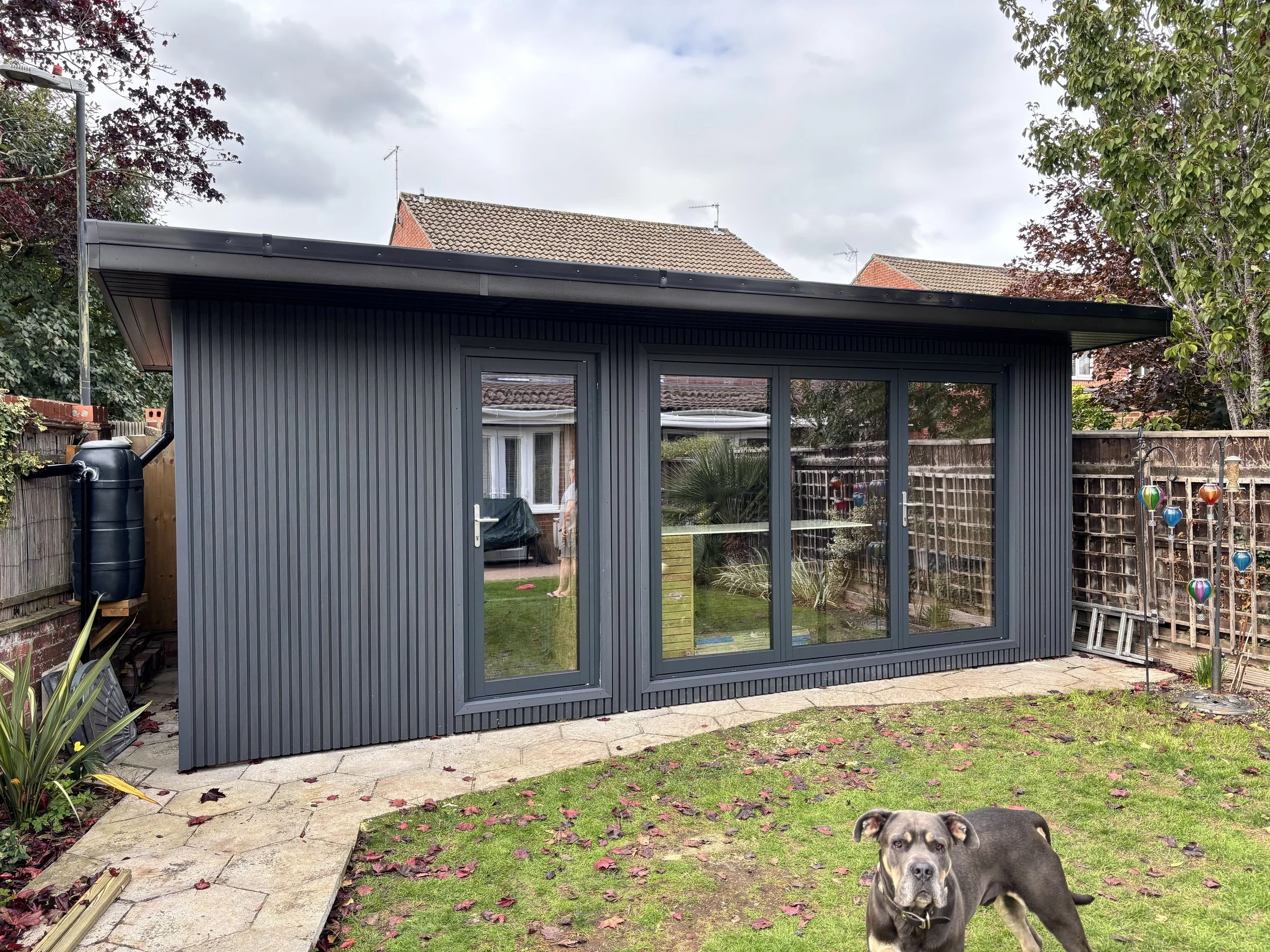 A small, modern backyard shed with gray corrugated metal siding, glass sliding doors, and a sloped roof. The shed is situated on a paved stone path in a backyard with grass, plants, and a cardboard cutout of a person reflected in the glass. There is a dog standing on the grass in the foreground, and a weather vane with colorfully painted glass ornaments on the right side of the yard.