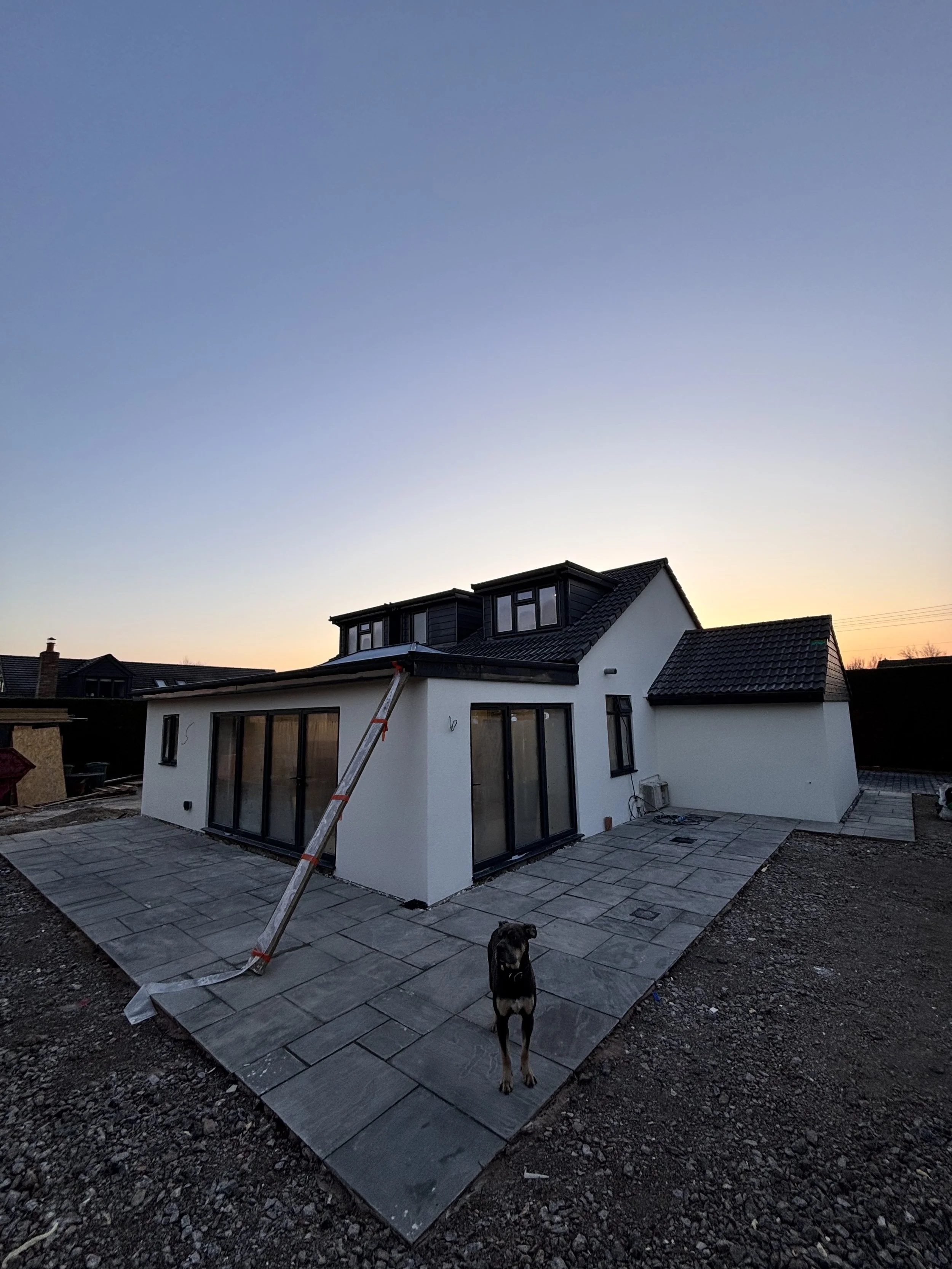 Newly constructed white house with large glass sliding doors, a small attached extension, and a paved patio area, with a dog standing on the patio during sunset.