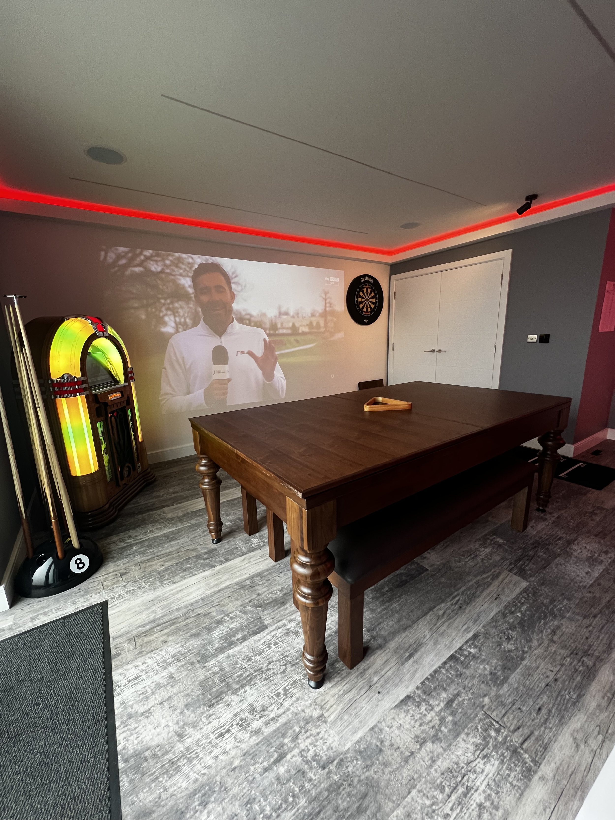 A room with a wooden table, a wall with a dartboard, a dart machine, and a projection of a man holding a microphone, standing outdoors.