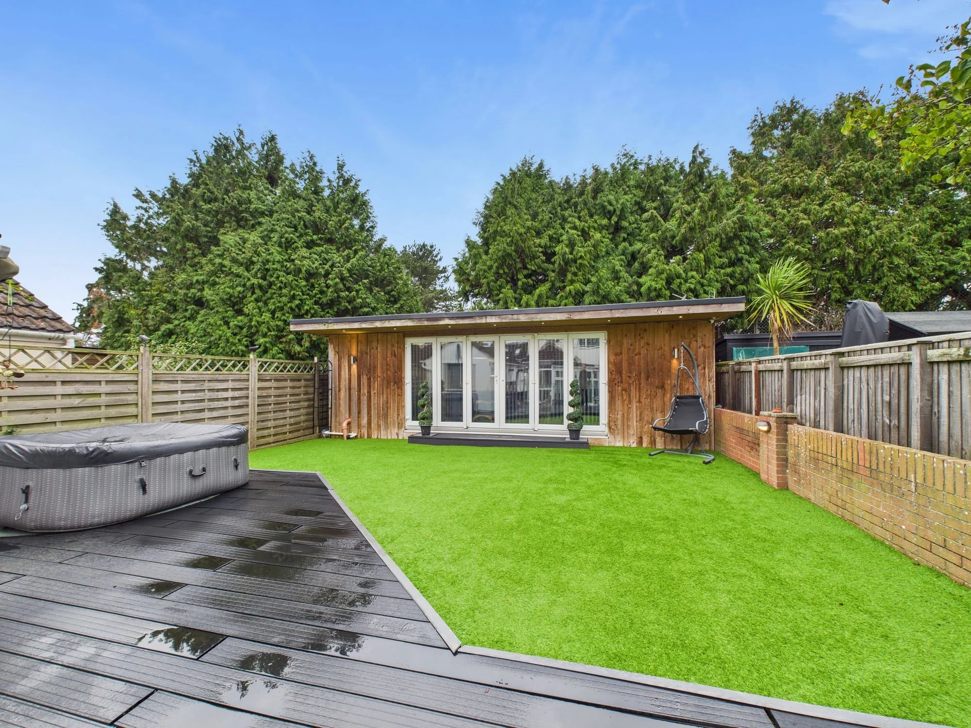 A backyard with a wooden shed, artificial grass, a hot tub, and a black swinging chair. There are tall trees and a fence surrounding the yard.