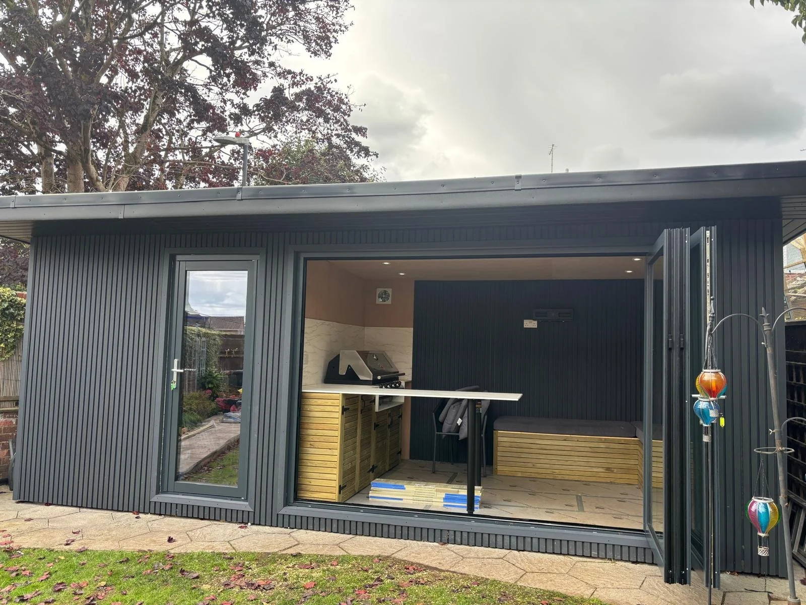 Backyard shed with open doors showing interior with a barbecue grill, a bench, and a small table, surrounded by trees and cloudy sky.