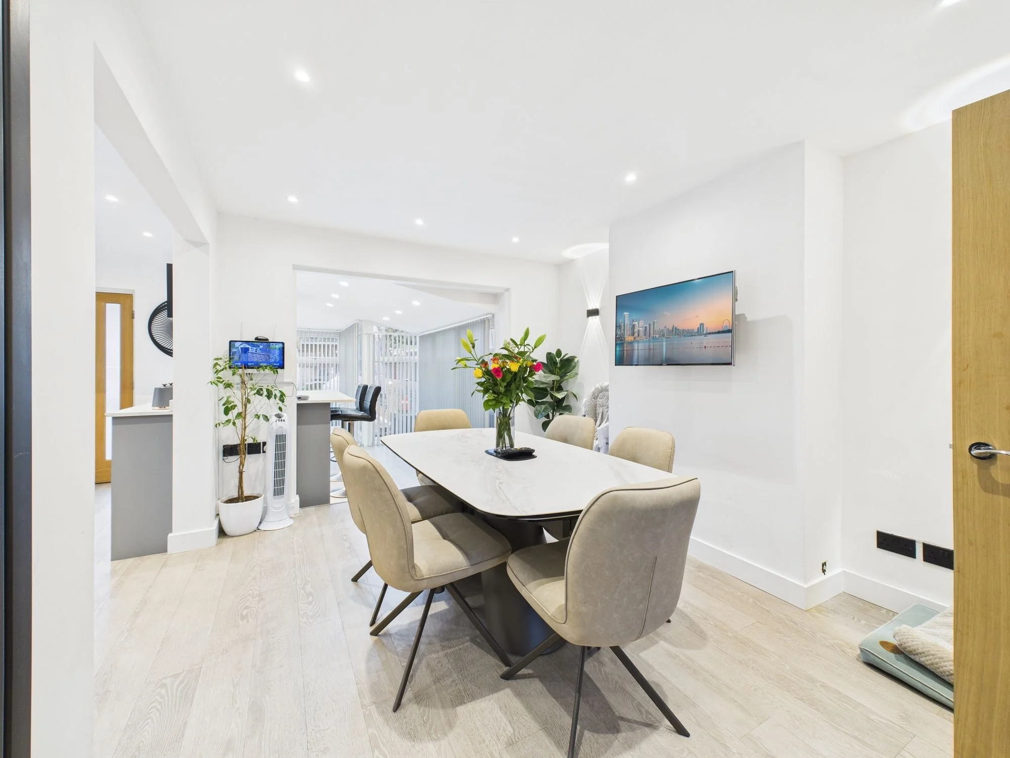 Modern dining room with a rectangular marble table, six beige upholstered chairs, a vase of flowers, wall-mounted TV, and bright natural light.