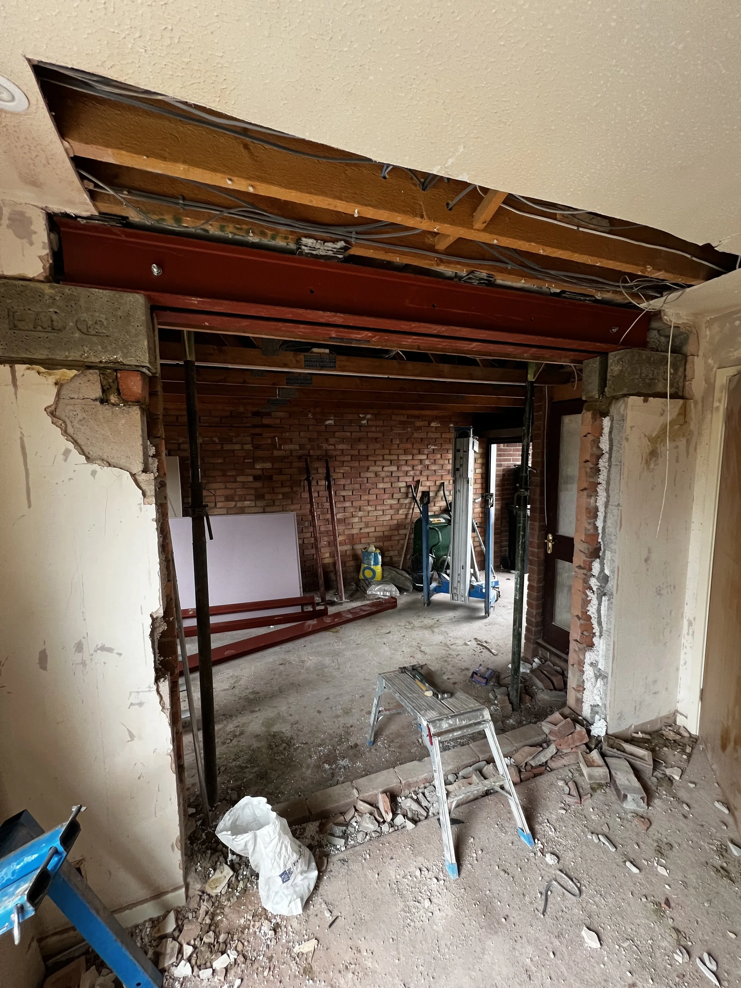 Interior wall demolition in a home renovation project, exposing brick wall and ceiling framing with construction tools and debris on the floor.