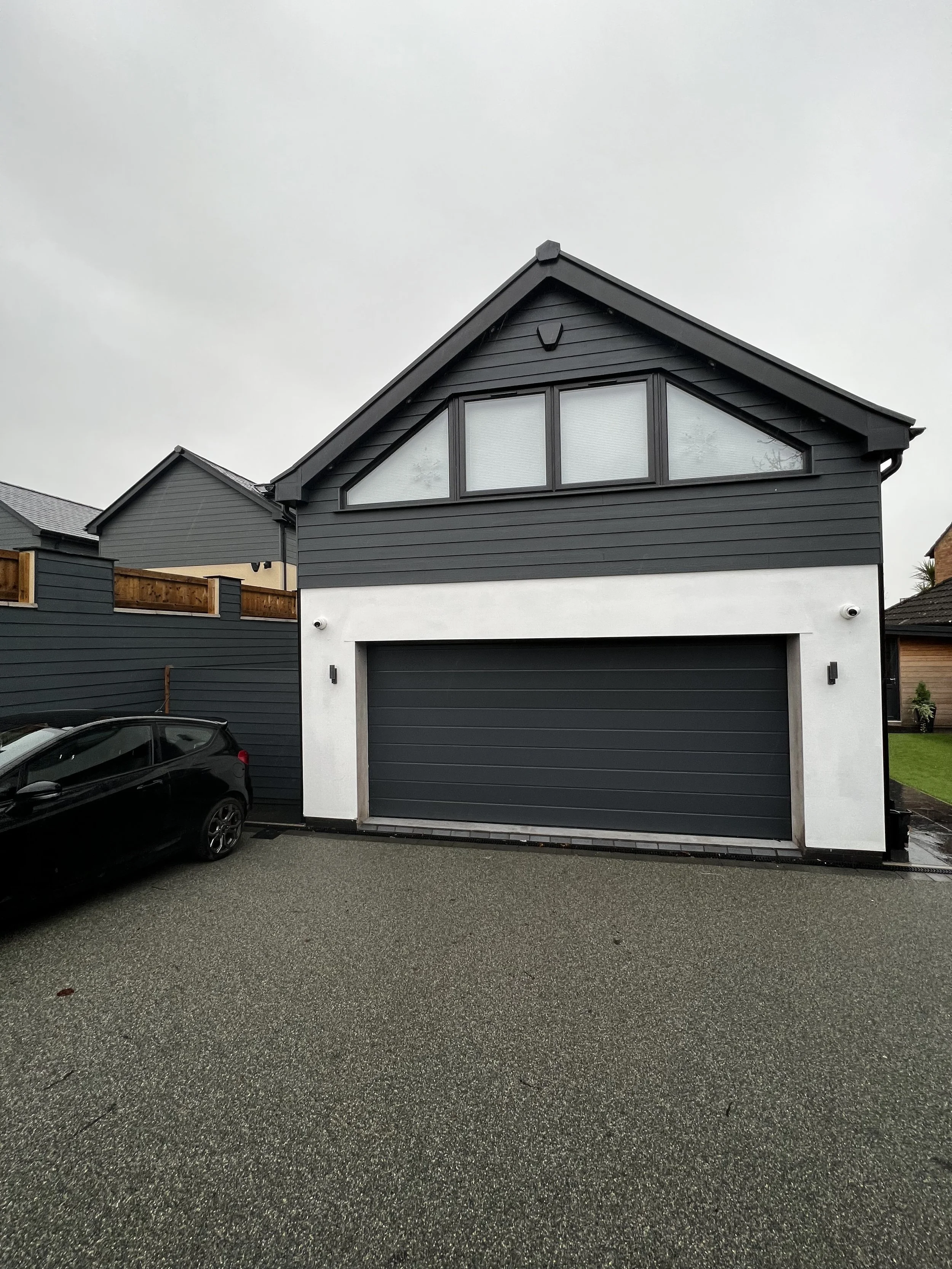 A modern two-story house with black siding, a large garage door, and triangular window above the garage, with a gray driveway and a black car parked on the side.