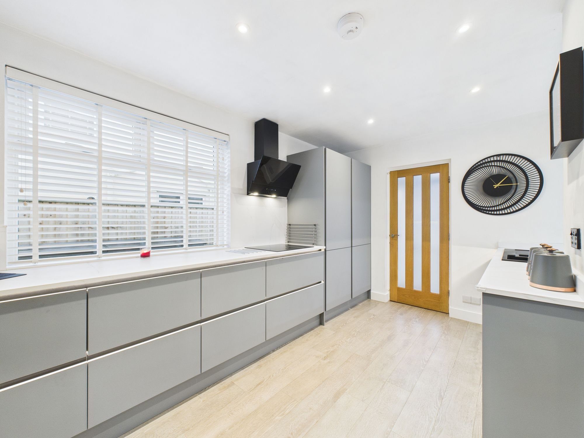 Modern kitchen with white countertops, gray cabinets, black range hood, large window with white blinds, wooden door, wall clock, and small appliances.