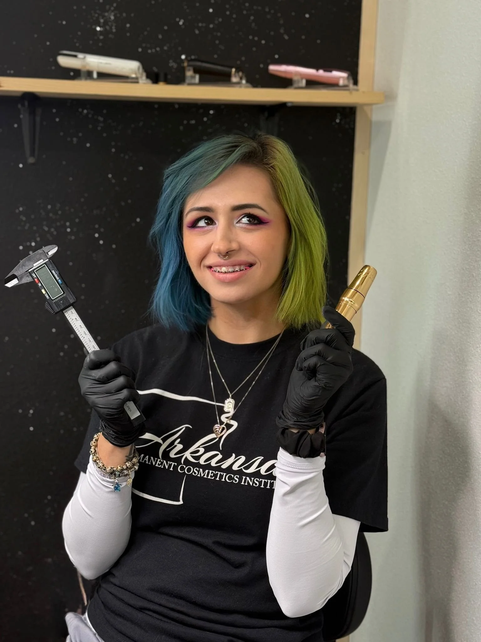 Tabitha Campbell, wearing black gloves, holding a digital caliper in one hand and a gold tattoo machine in the other, smiling with braces, in a studio with a black speckled background and shelves with makeup products.
