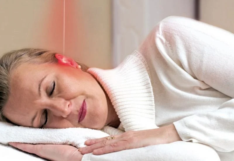 A woman with blonde hair and pink earrings is lying on her side, resting her head on a white pillow, smiling softly.