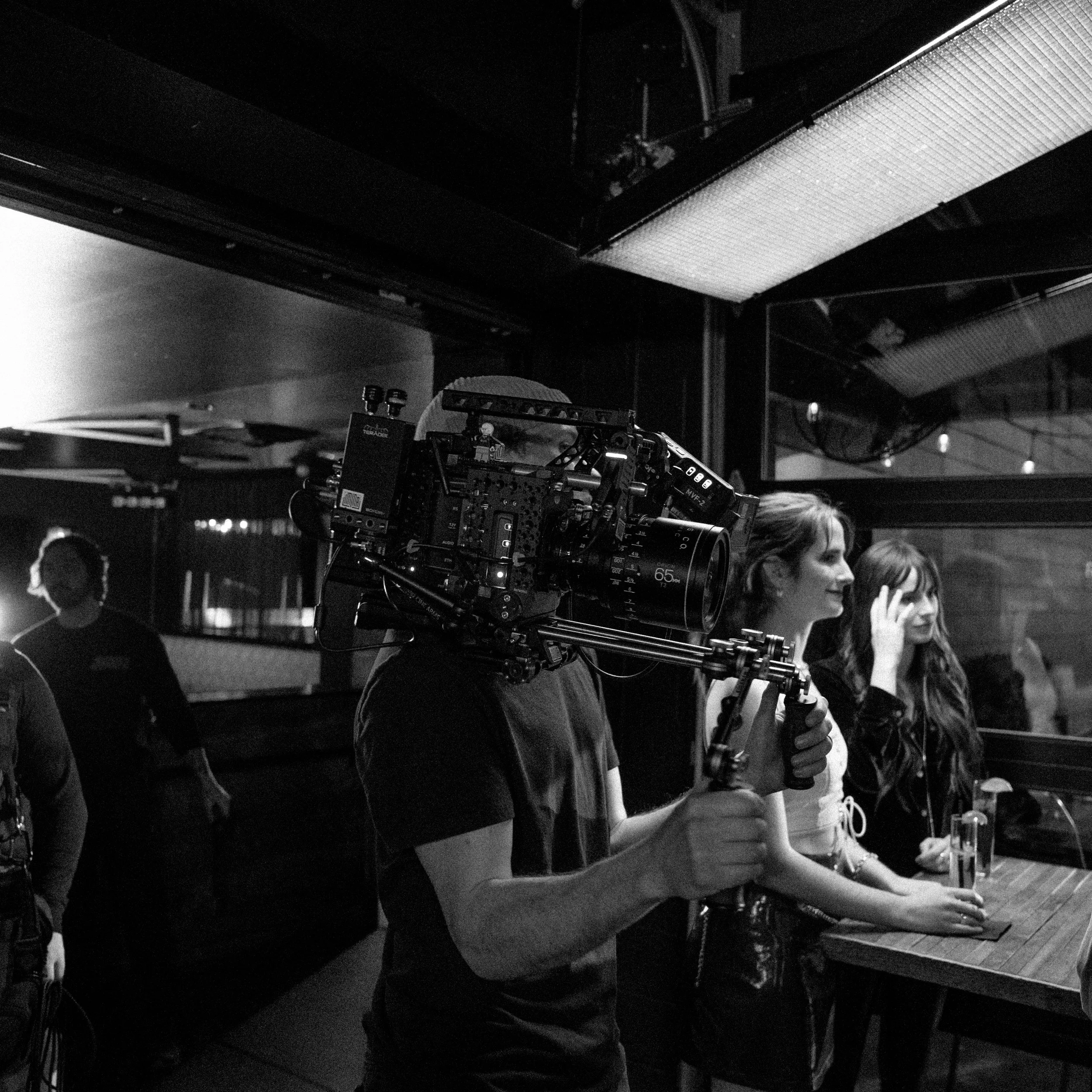 A person holding a professional camera filming two women sitting at a bar, with other people in the background at a lively indoor event.