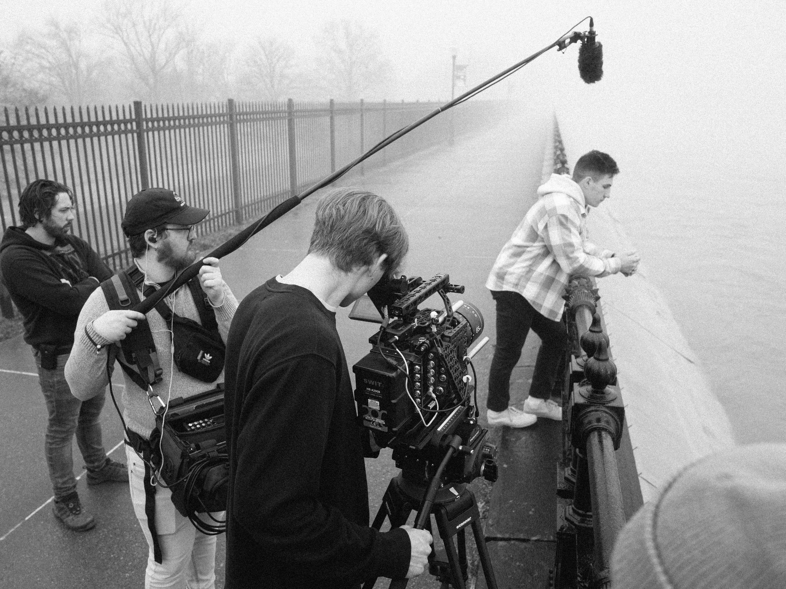 Filmmakers filming a scene by the water on a rainy day, with a young man leaning on the railing and a crew member operating a camera.