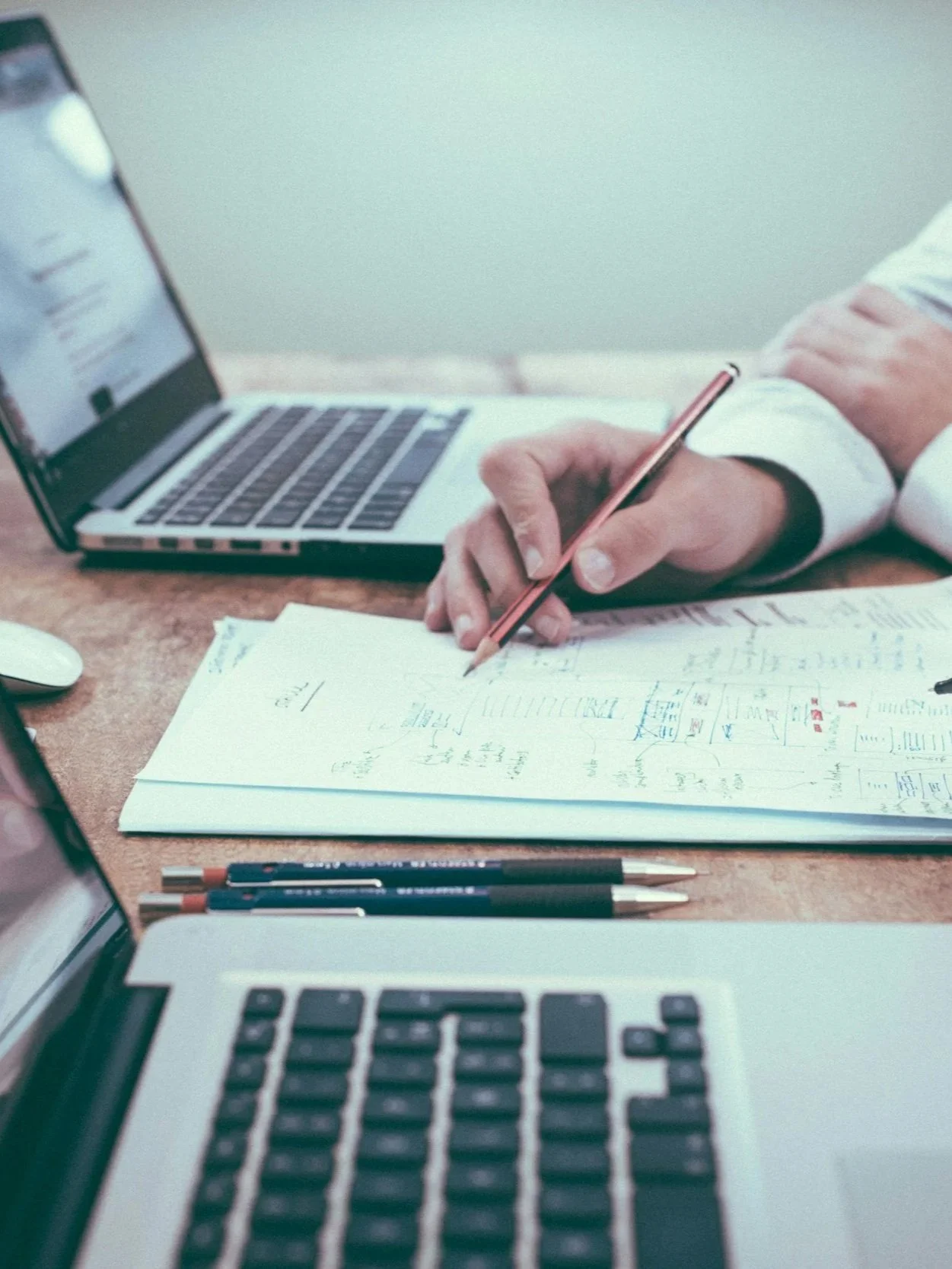 Person working at a desk with open laptops, notebooks, and pens, writing on papers.