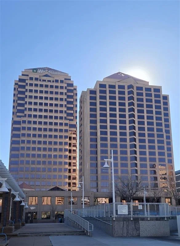 Two tall modern office buildings with glass windows, the one on the left has a sign for Wells Fargo bank at the top, under a clear blue sky with the sun shining behind the right building. Downtown Albuquerque buildings.
