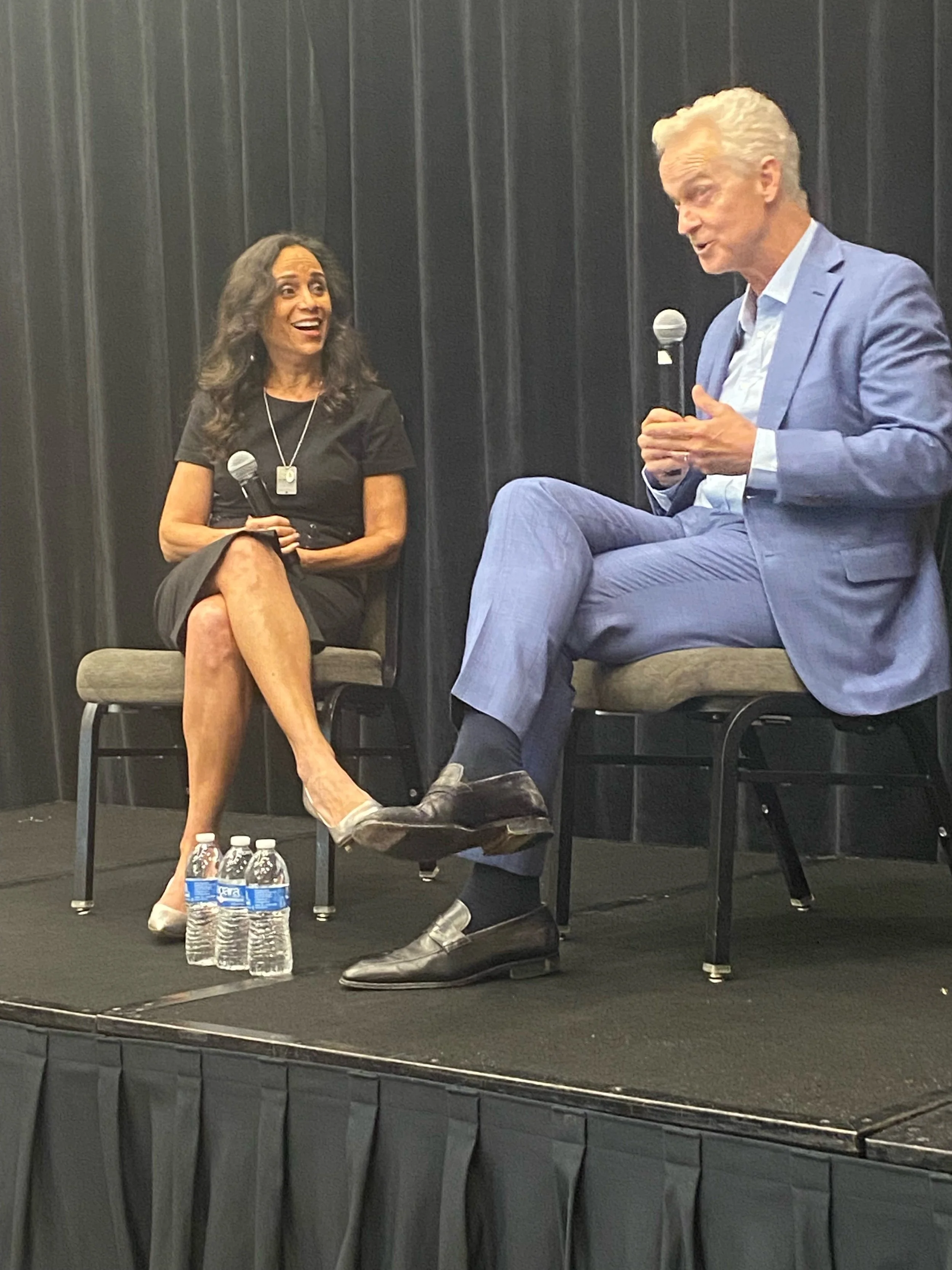 Two people, a woman and a man, seated on stage having a discussion. The woman is holding a microphone, wearing a black dress, and smiling. The man is in a light blue suit, holding a microphone, and speaking. There are three water bottles on the floor