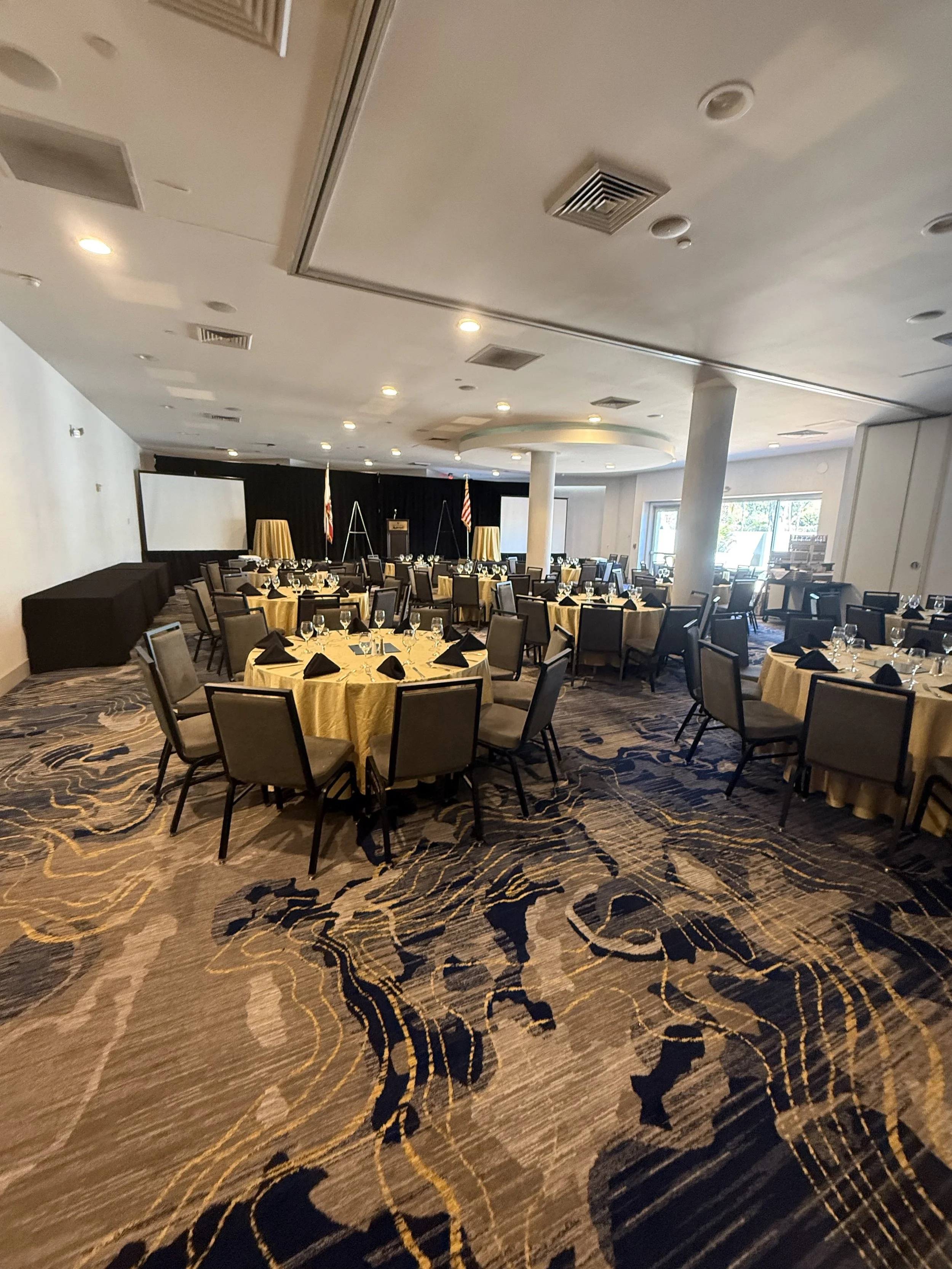 A banquet hall with round tables covered in yellow tablecloths, set with glassware, black napkins, and chairs around each table. A stage with a podium, American flags, and large projection screens is visible at the front. The room has a patterned carpet, white walls, and large windows on one side.