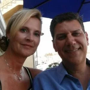 A smiling woman with blonde hair and a man with dark hair taking a selfie together outdoors under an umbrella.