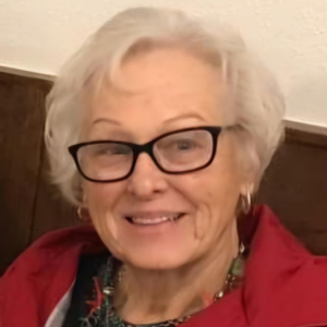 An elderly woman with white hair, wearing glasses and a red jacket, smiling at the camera.