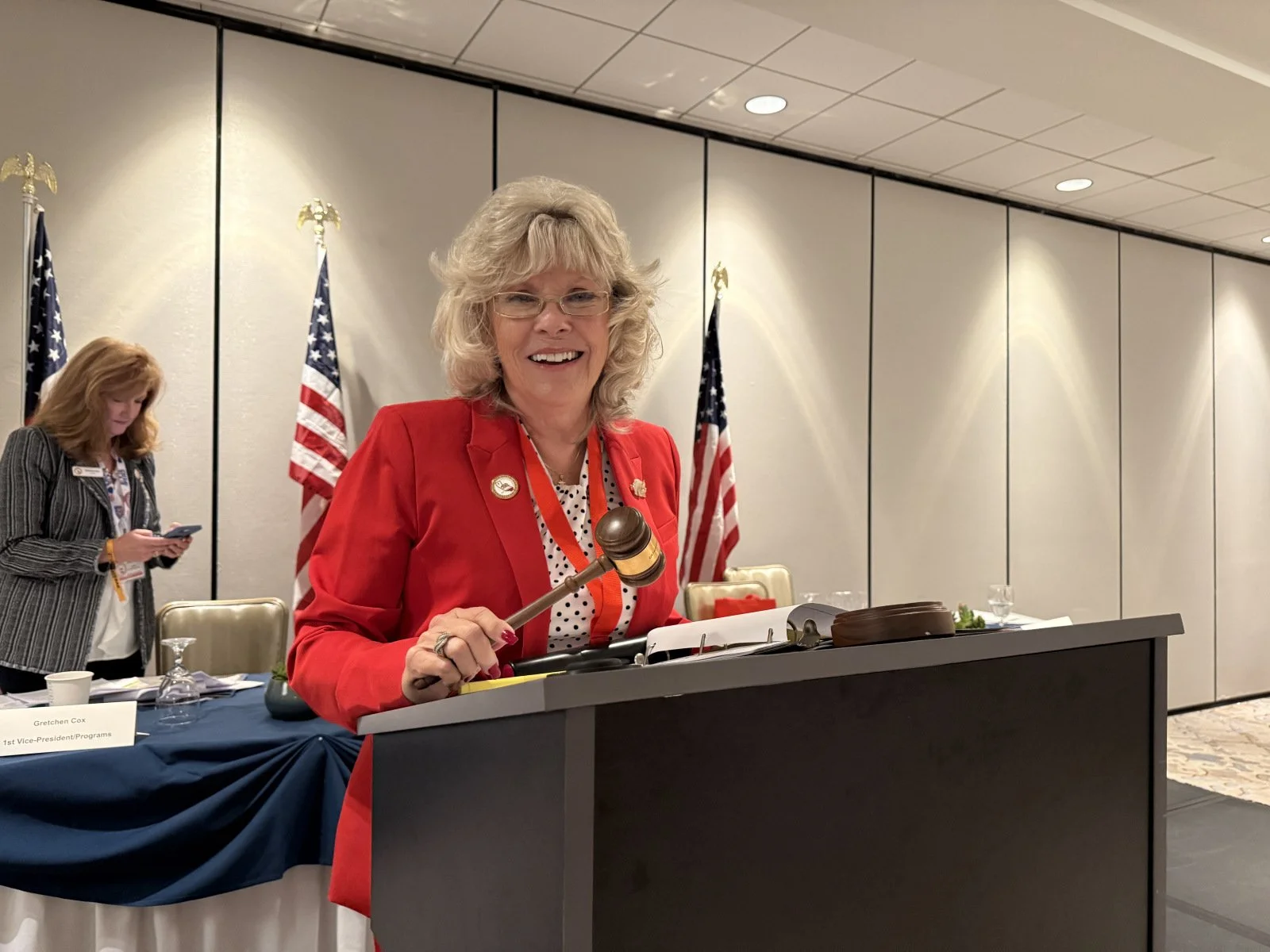 A woman in a red blazer holding a gavel, smiling, at a formal event with American flags behind her.