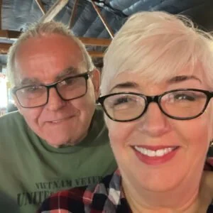 Close-up of a smiling man and woman wearing glasses, taking a selfie together indoors.