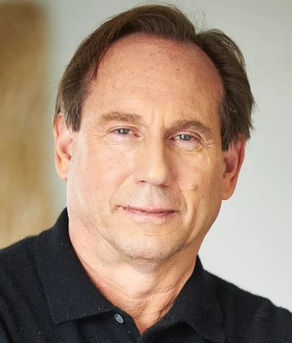 Close-up of a middle-aged man with brown hair, wearing a black polo shirt, in an indoor setting.