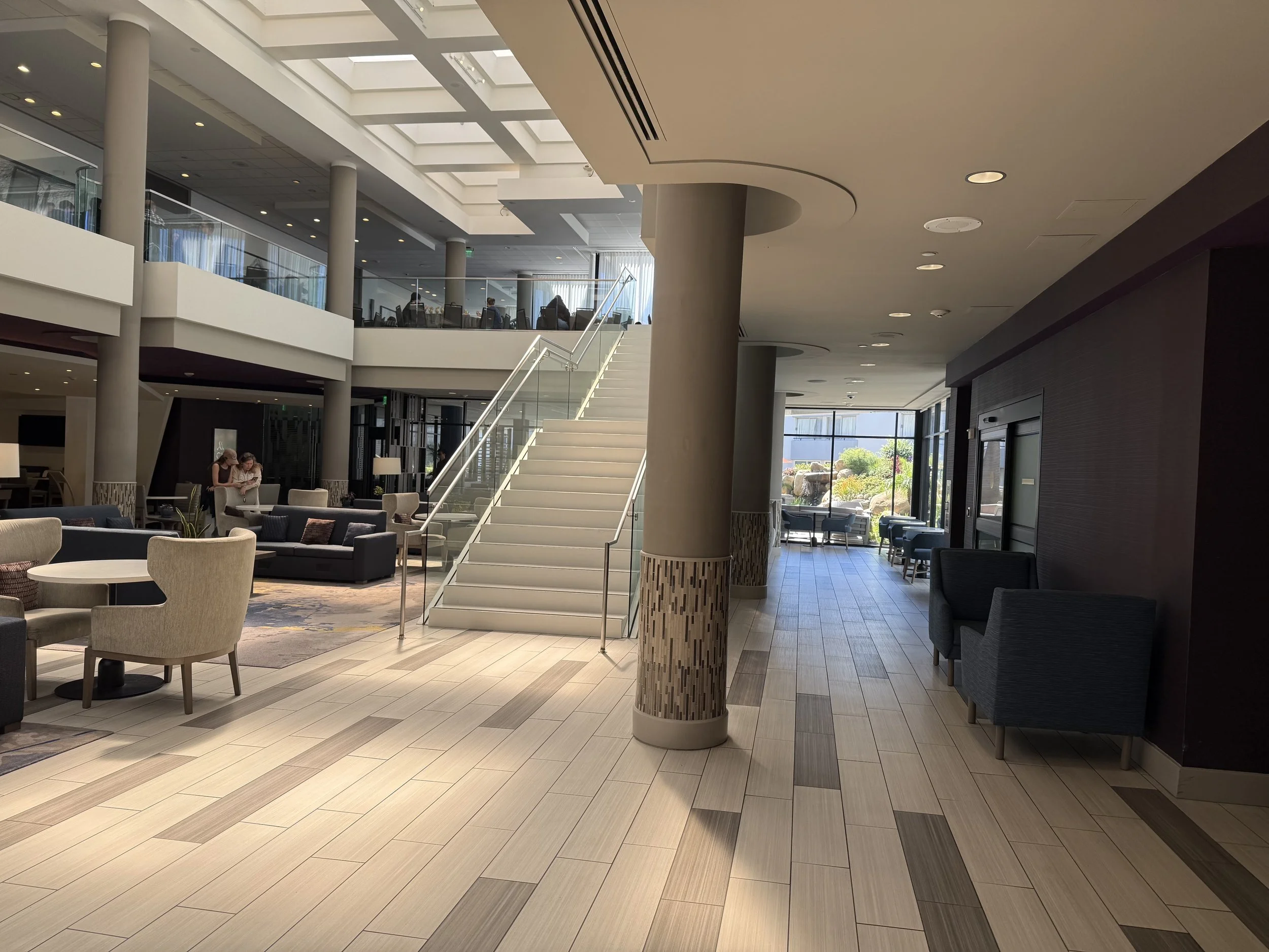 Interior of a modern hotel lobby with seating areas, a staircase, and large windows letting in natural light.