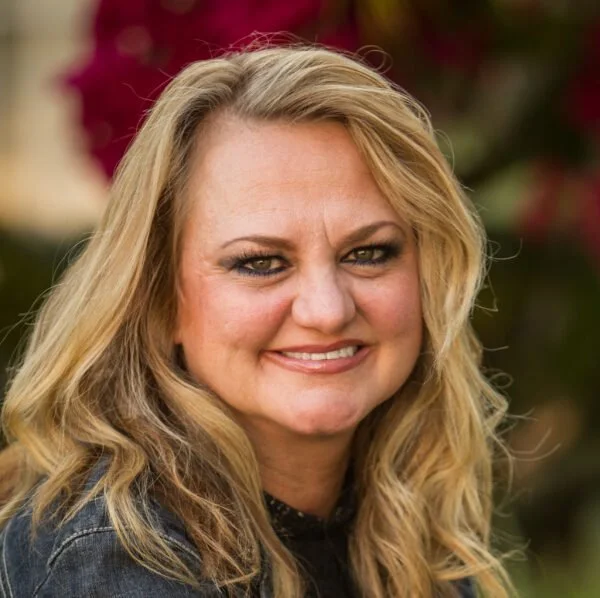 A smiling blonde woman with wavy hair, wearing a dark top, outdoors with pink and green foliage in the background.