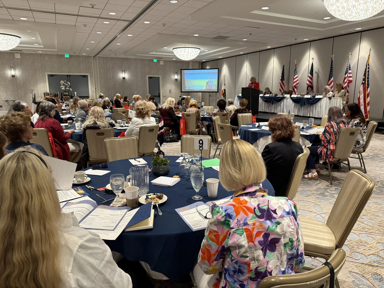 A conference or seminar room filled with women seated at round tables, listening to a speaker at a podium. The room is decorated with American flags and a large screen displaying a presentation.