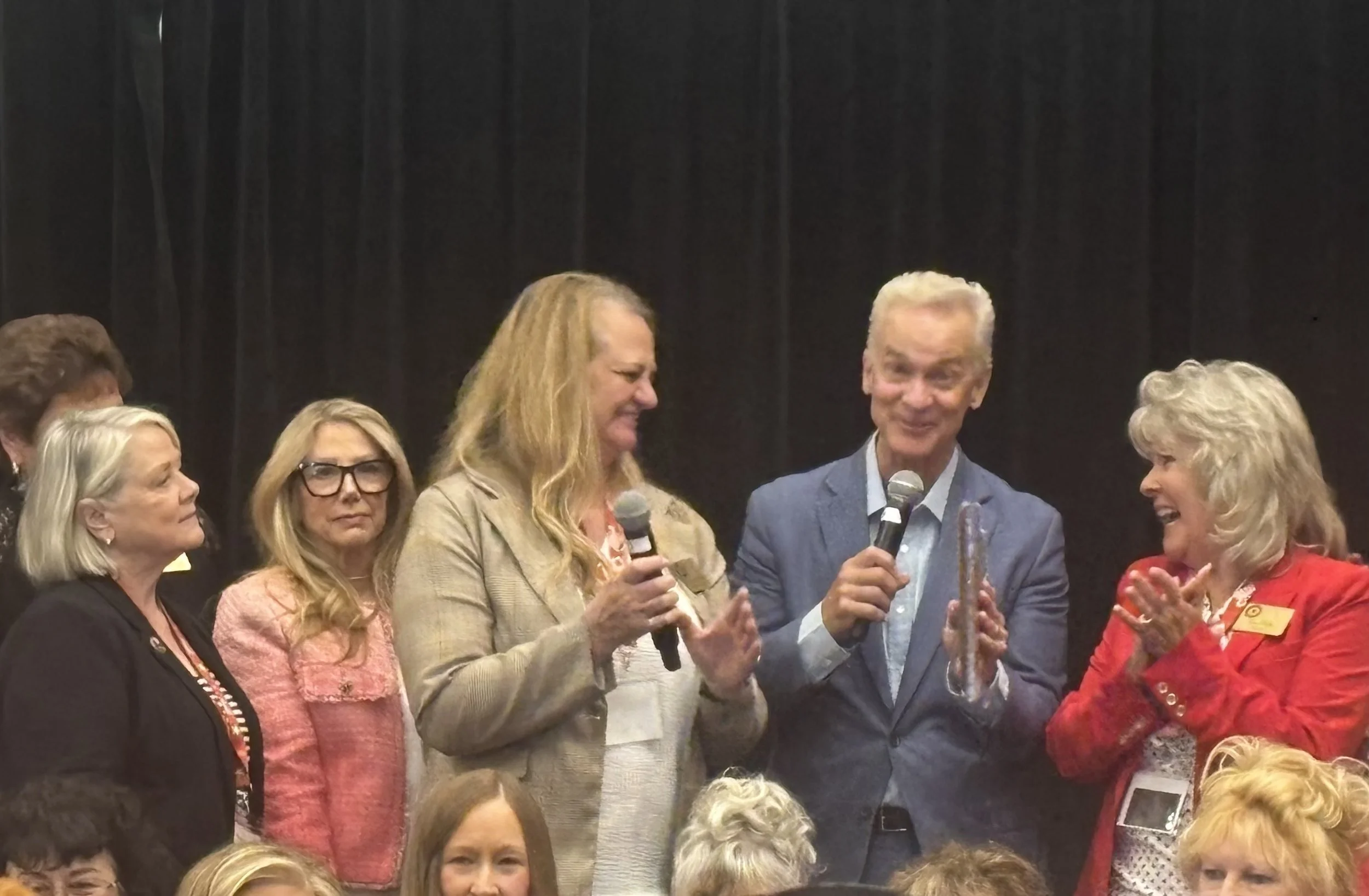 A group of women and one man on stage, with some holding microphones, in front of a black curtain, engaging in conversation or presentation. The women are dressed in various colors and styles, and the man is wearing a blue blazer.