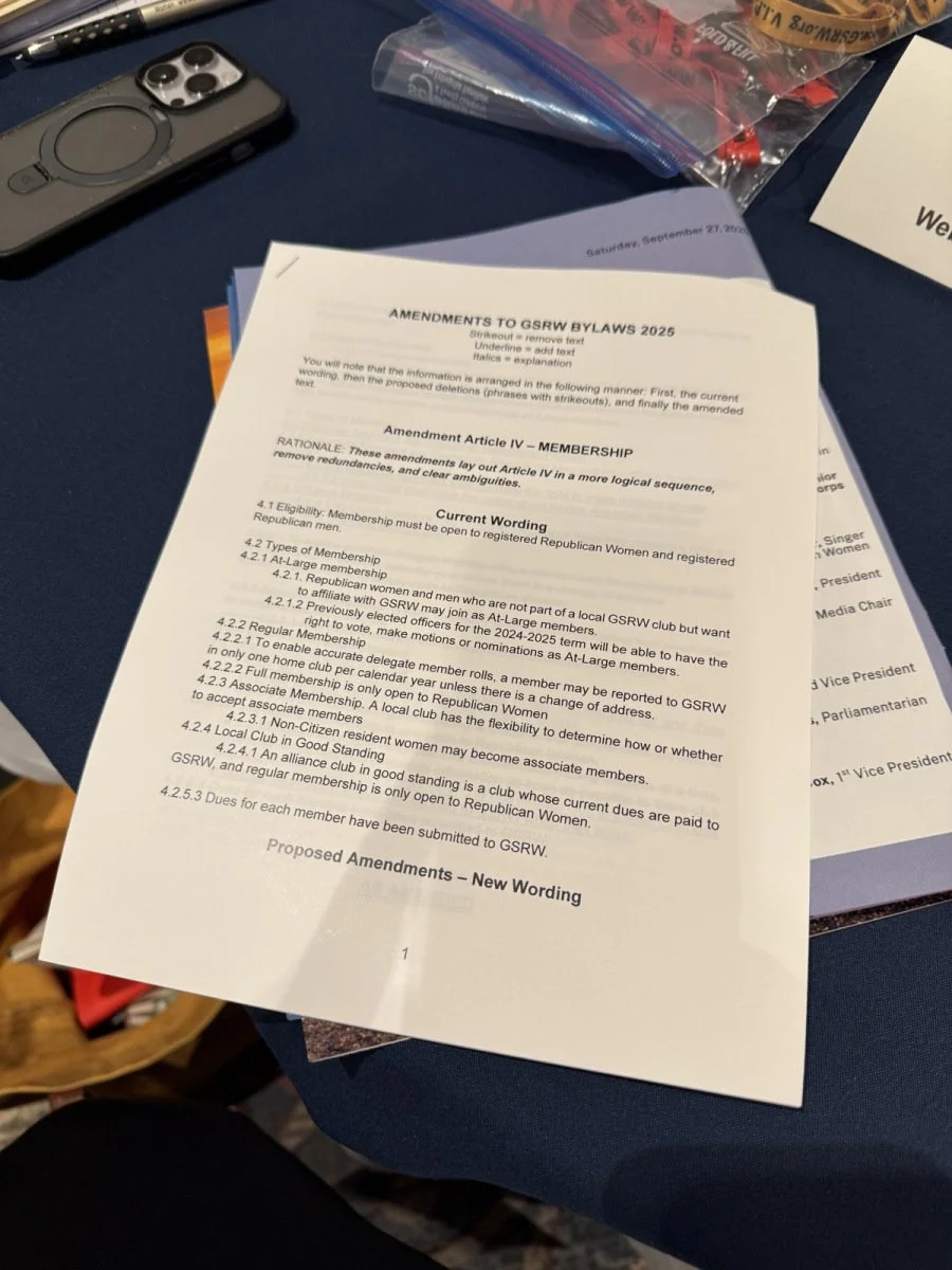 A printed document titled 'Amendments to GSRW Bylaws 2025' is placed on a dark blue table, with a smartphone, some pens, and a plastic bag with assorted items nearby.